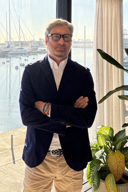 Hombre con gafas y chaqueta azul cruza brazos frente a ventana con vista al puerto con barcos y plantas.