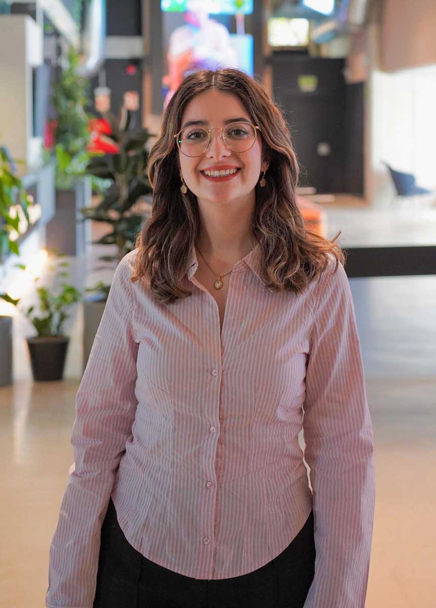 Mujer sonriente con gafas y blusa rosa de rayas en un espacio moderno con plantas.