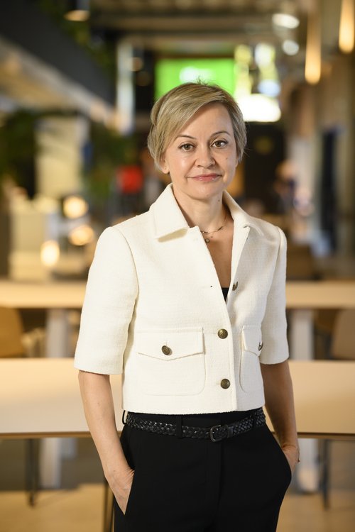 Mujer con chaqueta blanca y pantalones negros en un entorno interior moderno y bien iluminado.