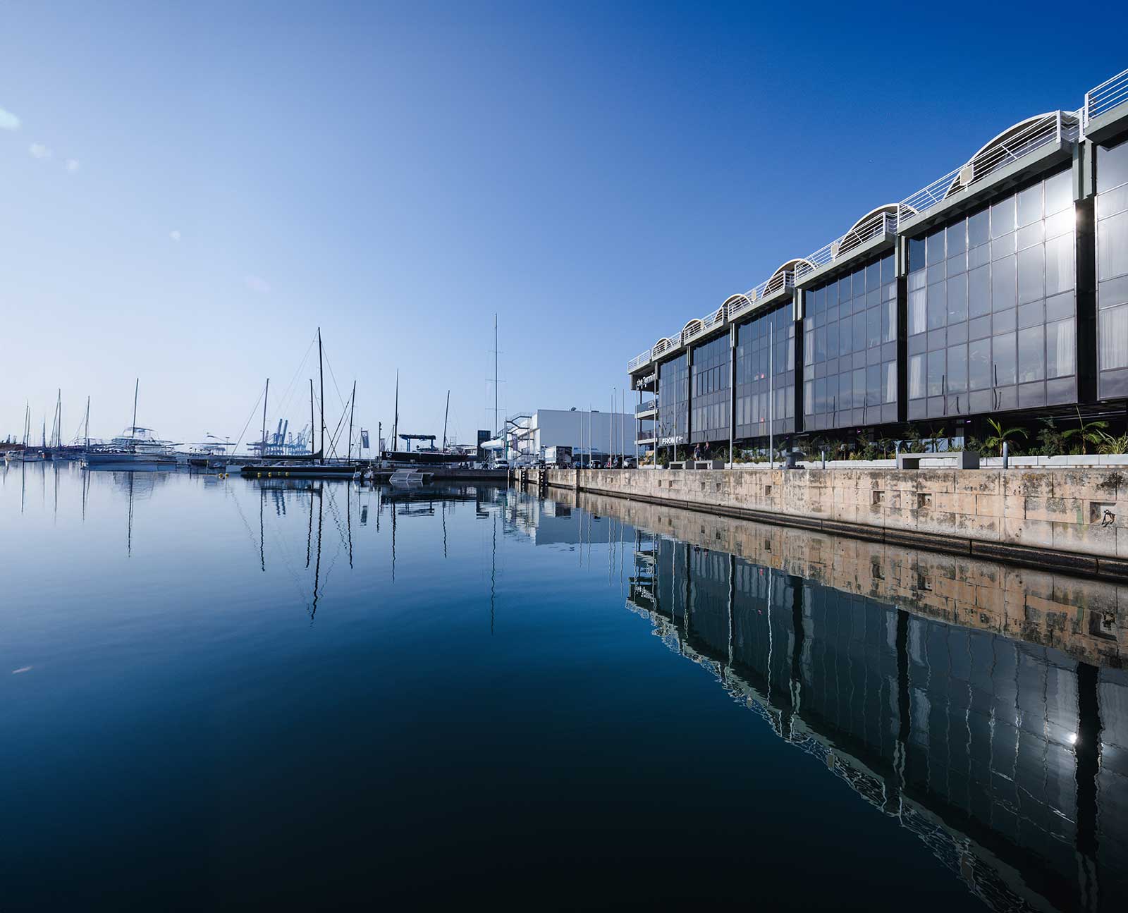 Puerto con embarcaciones y edificio moderno de cristal reflejado en agua tranquila bajo un cielo azul claro.
