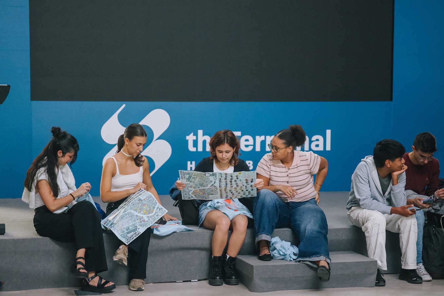 Seis jóvenes sentados con mapas y teléfonos frente a un fondo azul de the Terminal Hub.