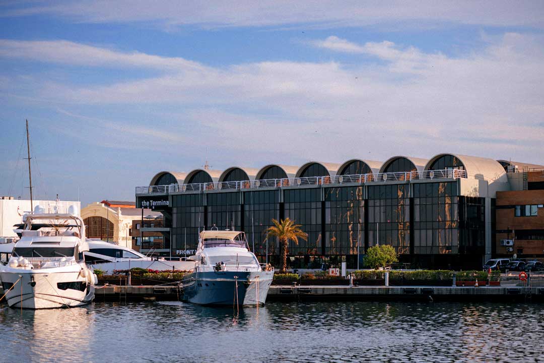 Edificio moderno con techo curvo junto a un muelle con varios yates amarrados en agua tranquila.
