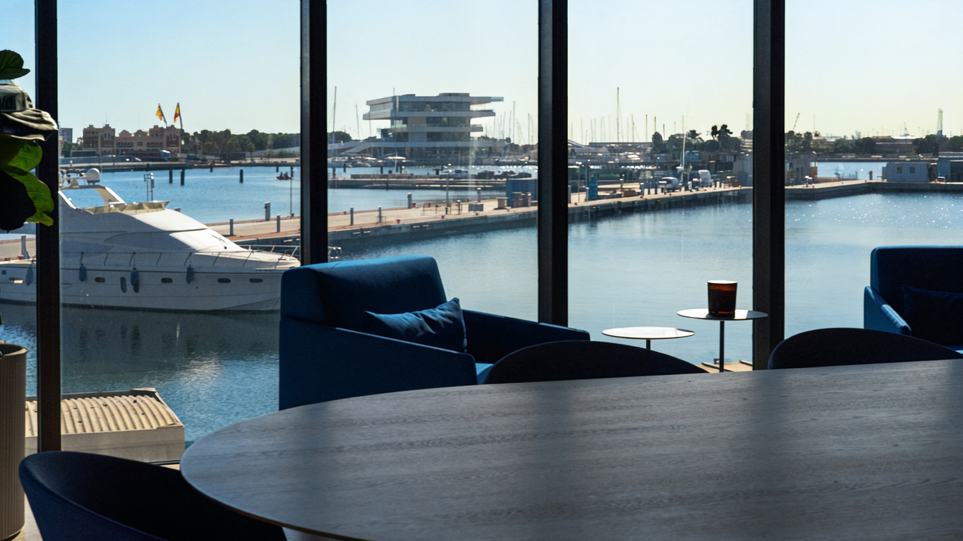 Sala con vista al puerto, sofá azul y mesas frente a ventanales con yate y agua en el fondo.