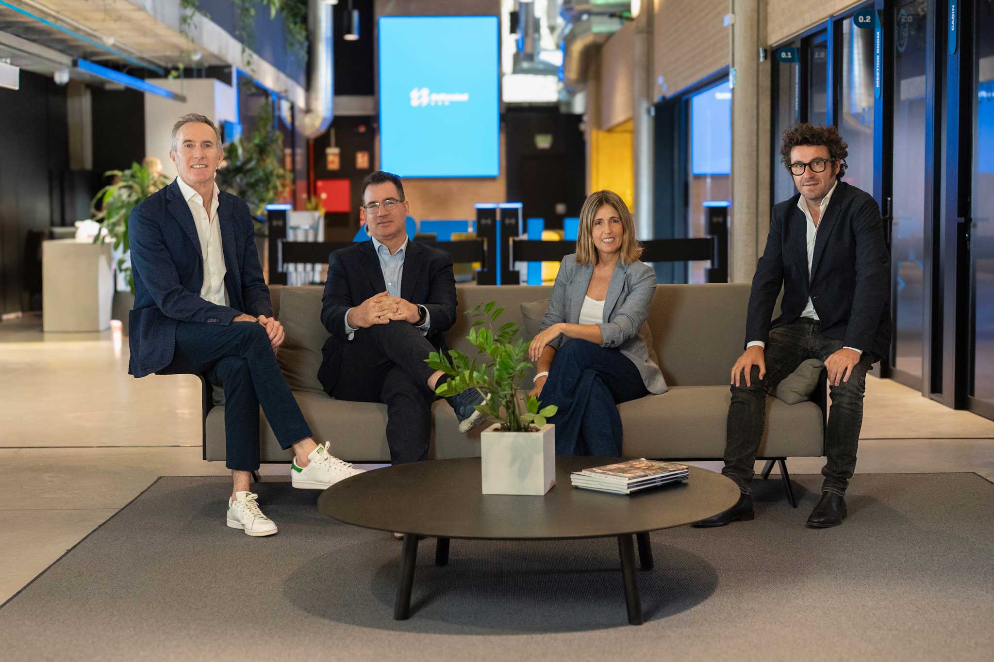 Cuatro personas sentadas en un sofá en un espacio moderno con pared de cristal y mesa redonda con planta.