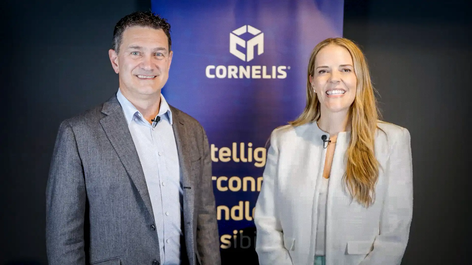 A man in a gray blazer and a woman in a light-colored jacket smiling, standing in front of a blue banner with the Cornelis logo.