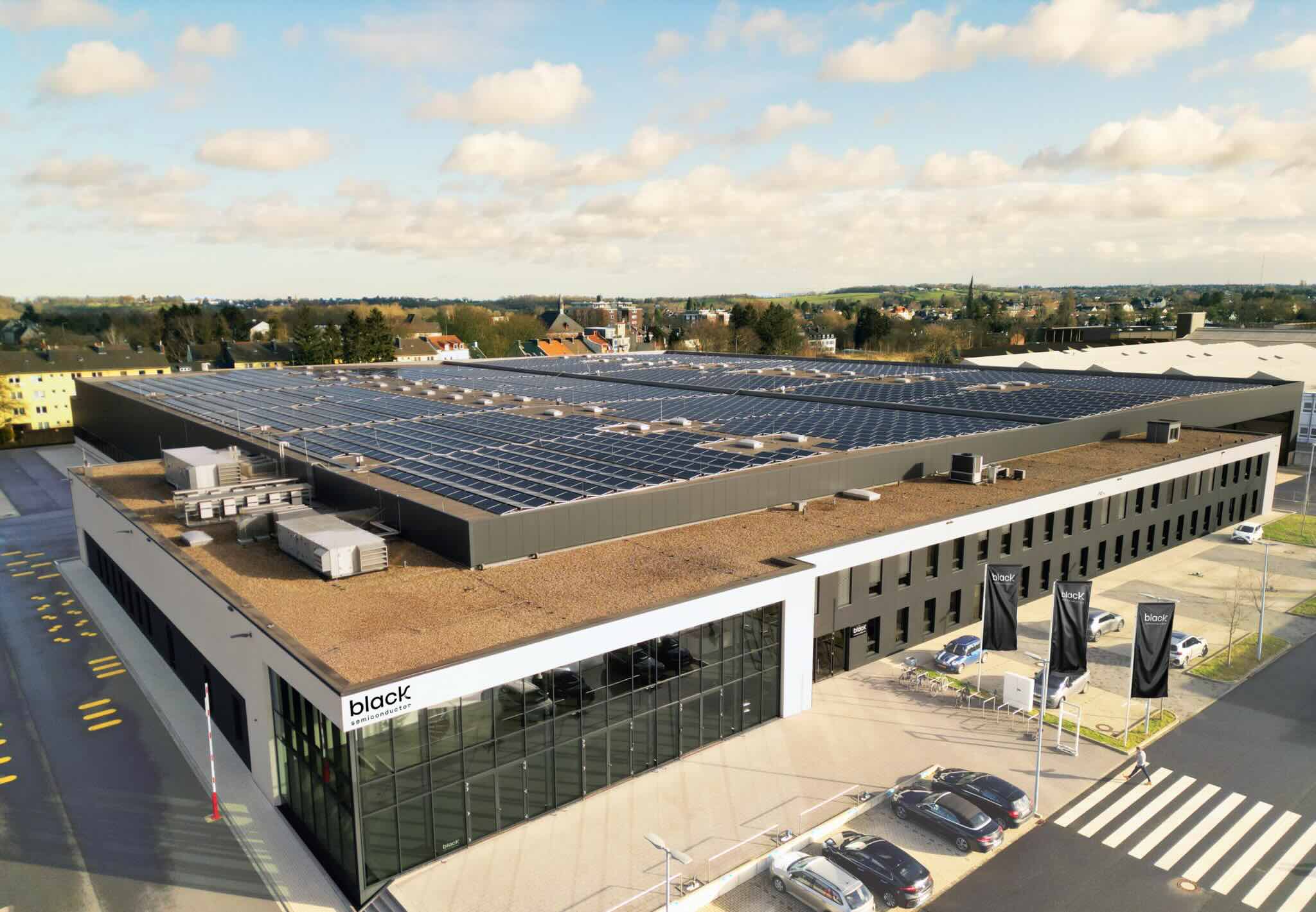 Modern industrial building with a large rooftop covered in solar panels and parked cars outside.