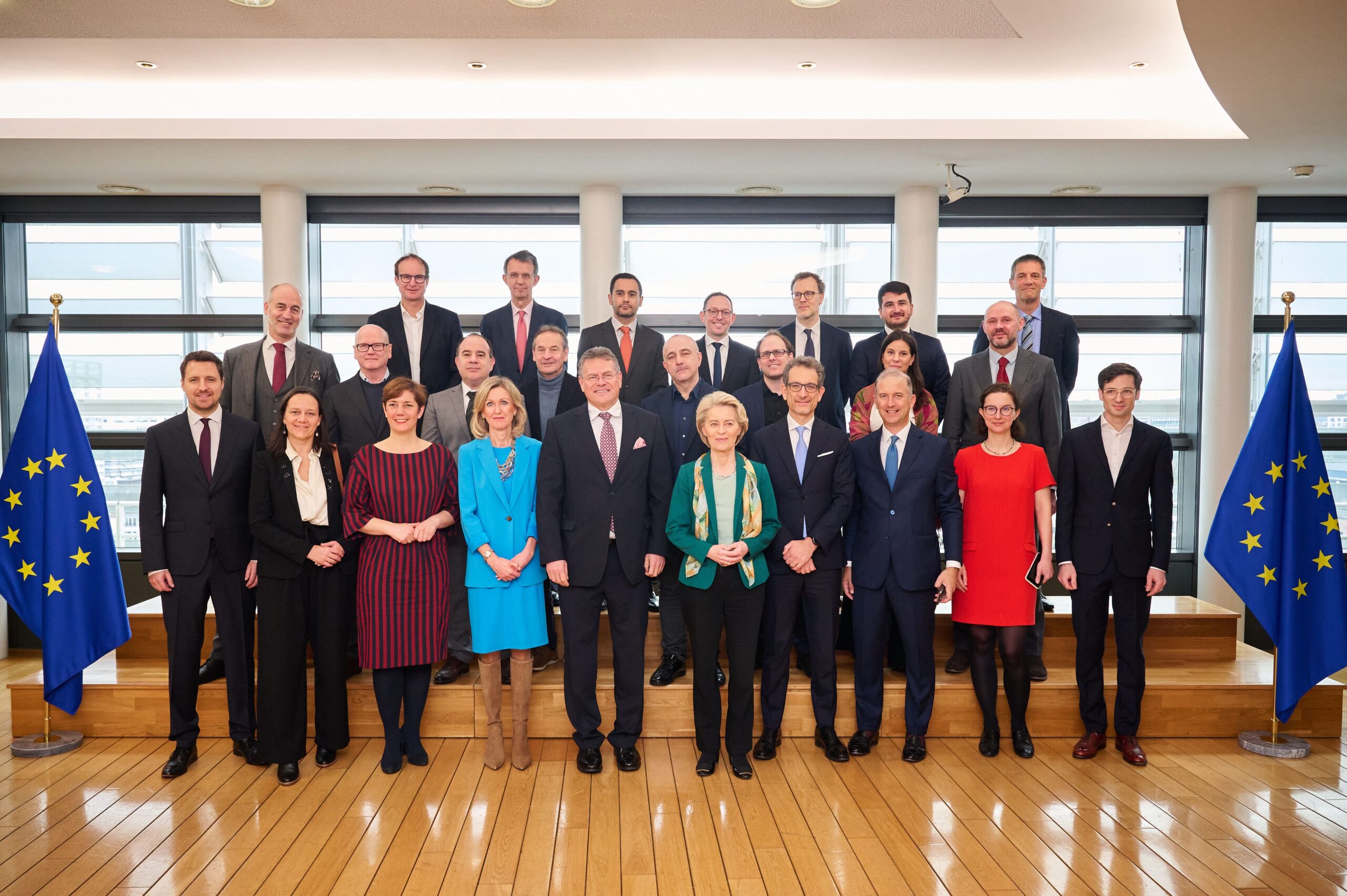 Group of 24 professionally dressed men and women posing inside a modern building with European Union flags on each side.