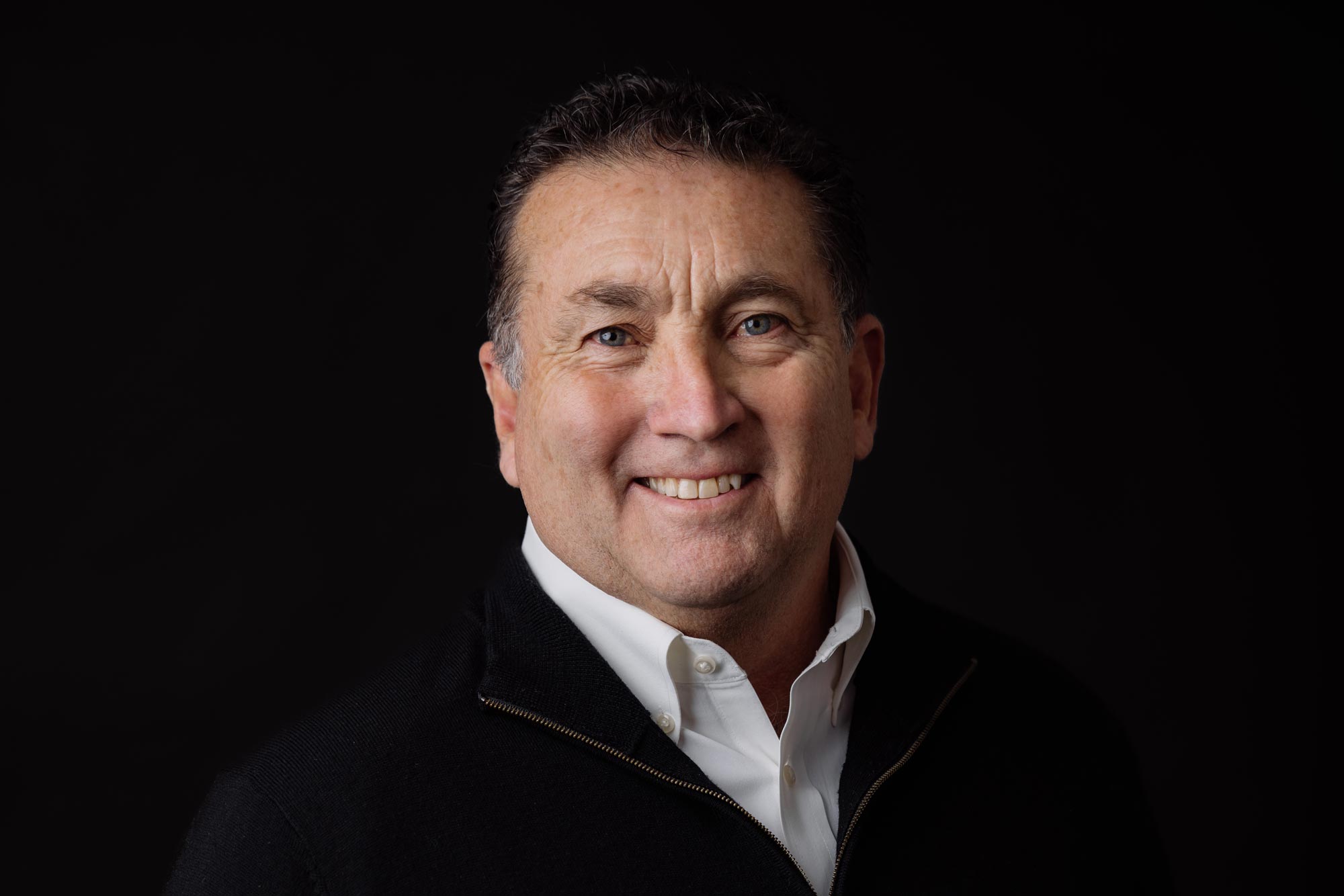 Smiling middle-aged man with short hair, wearing a white collared shirt and dark jacket against a dark background with green accent light.