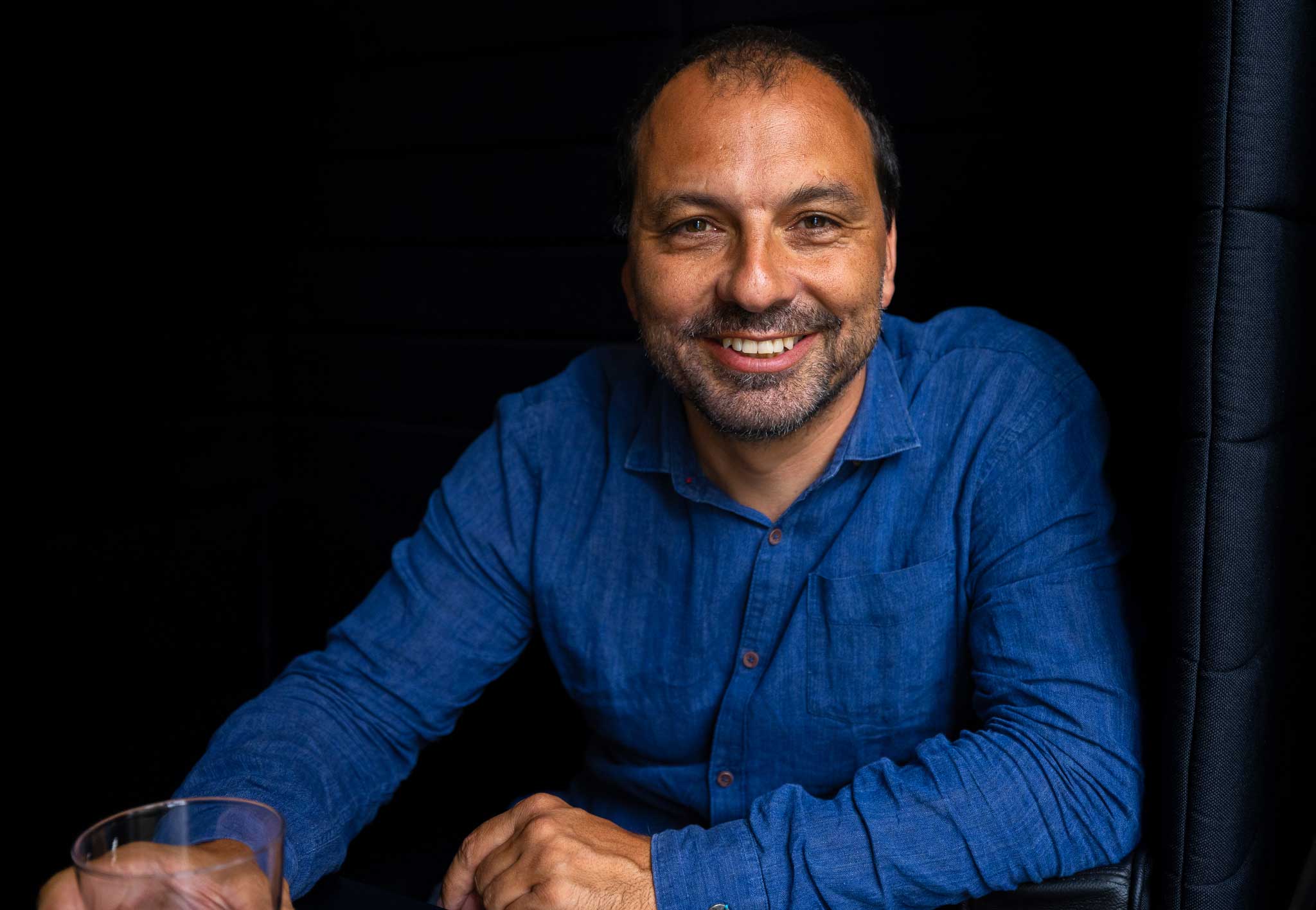 Smiling man in a blue shirt sitting against a black background holding a glass.