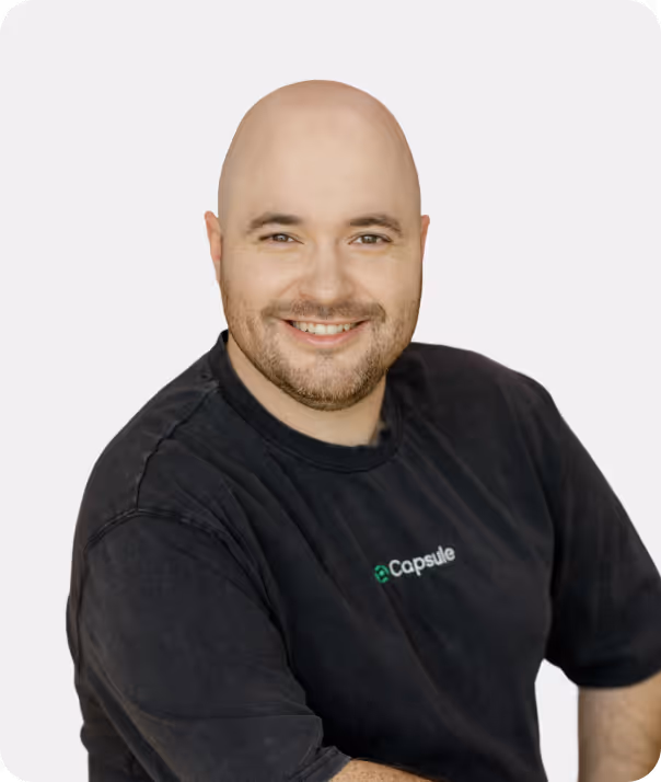 Smiling bald man with a short beard wearing a black Capsule t-shirt against a light background.