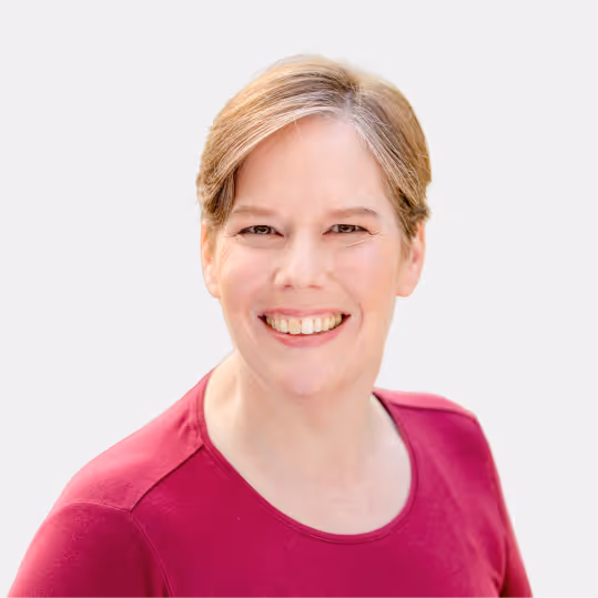 Smiling woman with short light brown hair wearing a dark pink shirt on a plain light background.