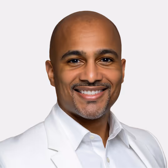 Smiling bald man with a trimmed beard wearing a white shirt and jacket against a light gray background.