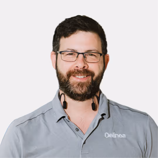 Smiling man with glasses and a beard wearing a gray Delinea polo shirt and black neckband headphones.