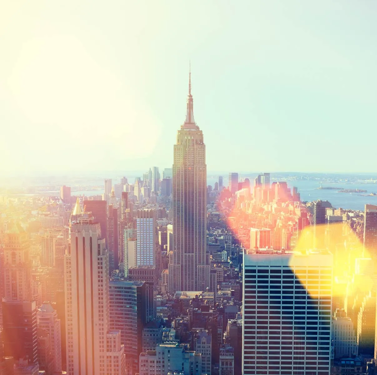 Sunlit view of the Empire State Building towering over New York City with a hazy skyline and yellow light flares.