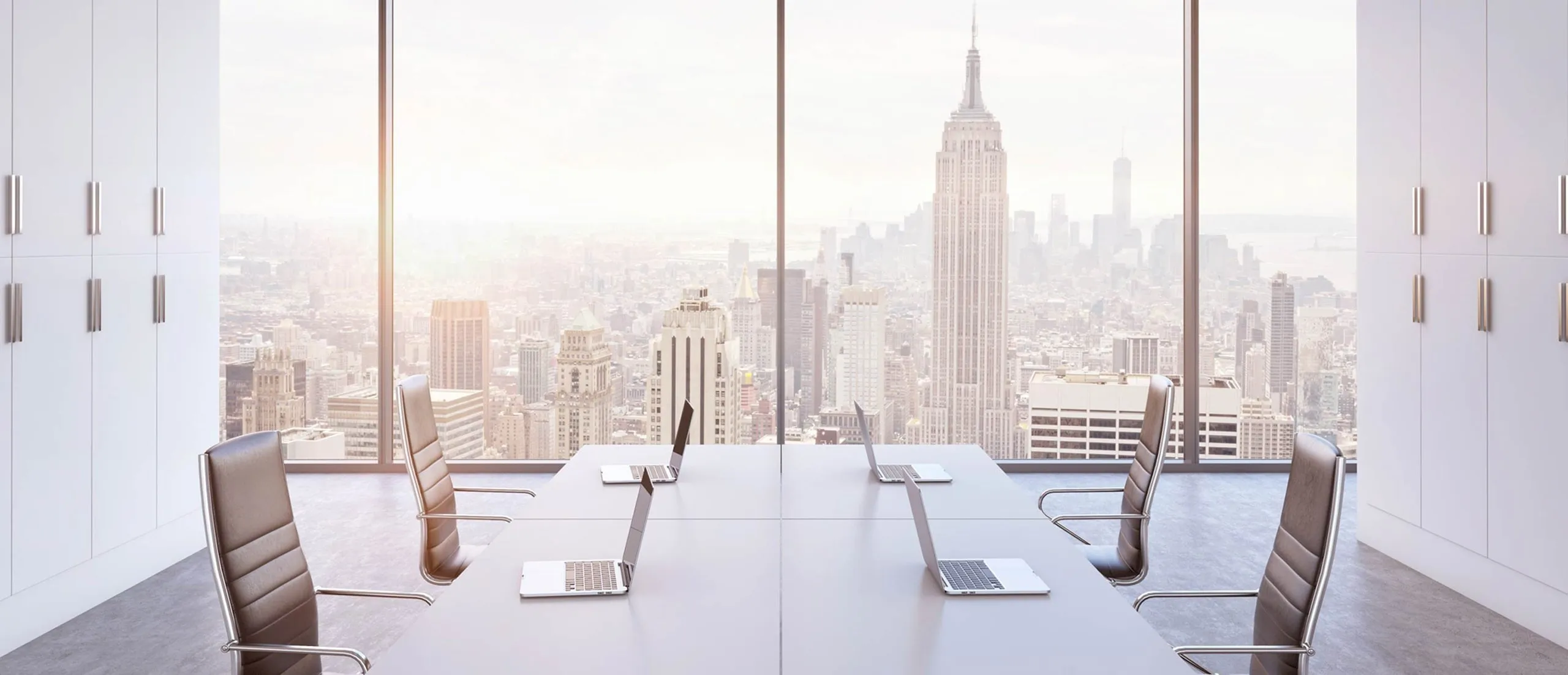 Modern conference room with six office chairs and four laptops on a long table, overlooking a city skyline with the Empire State Building.