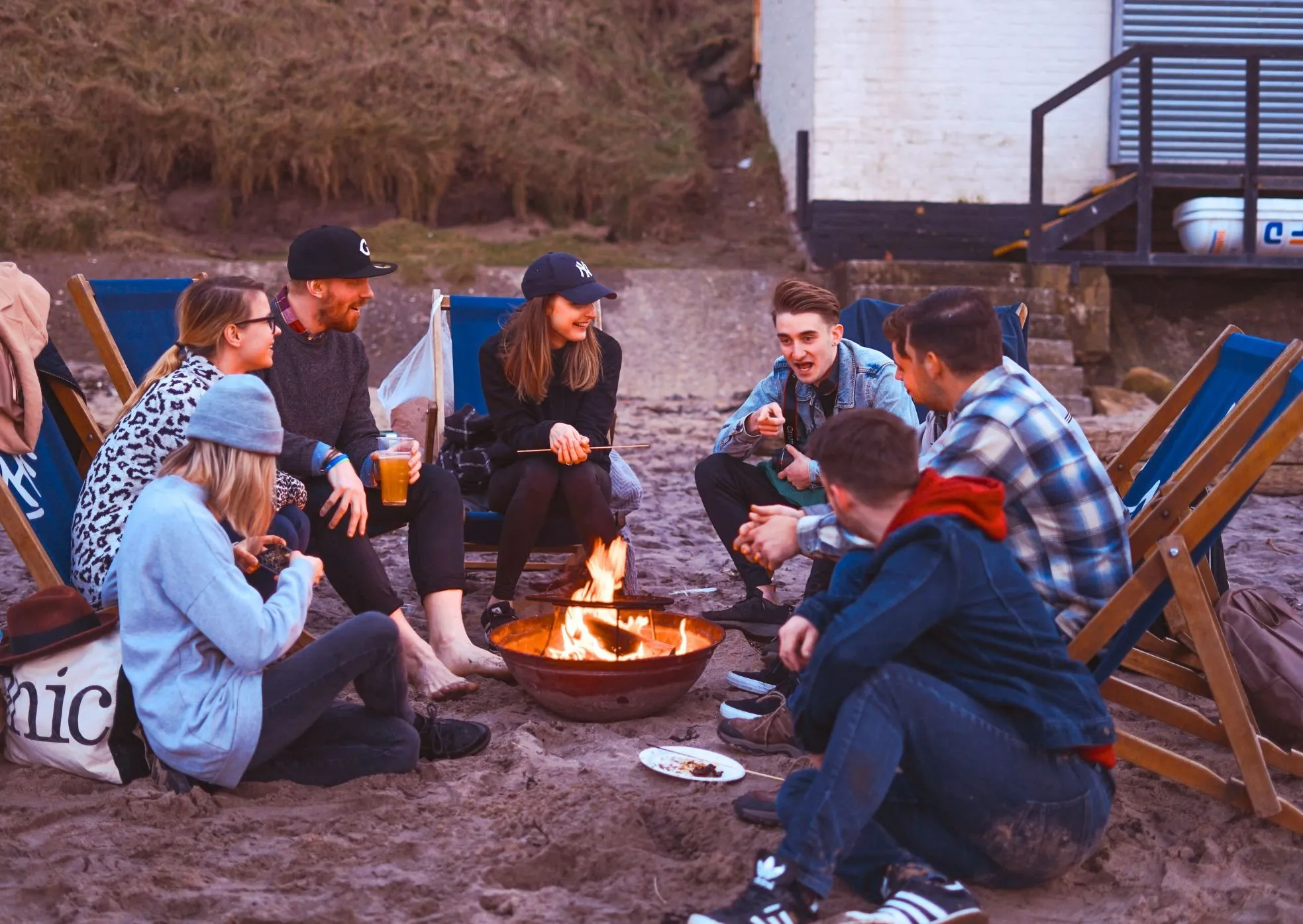 Group of young adults sitting on sand around a campfire, talking and roasting marshmallows.