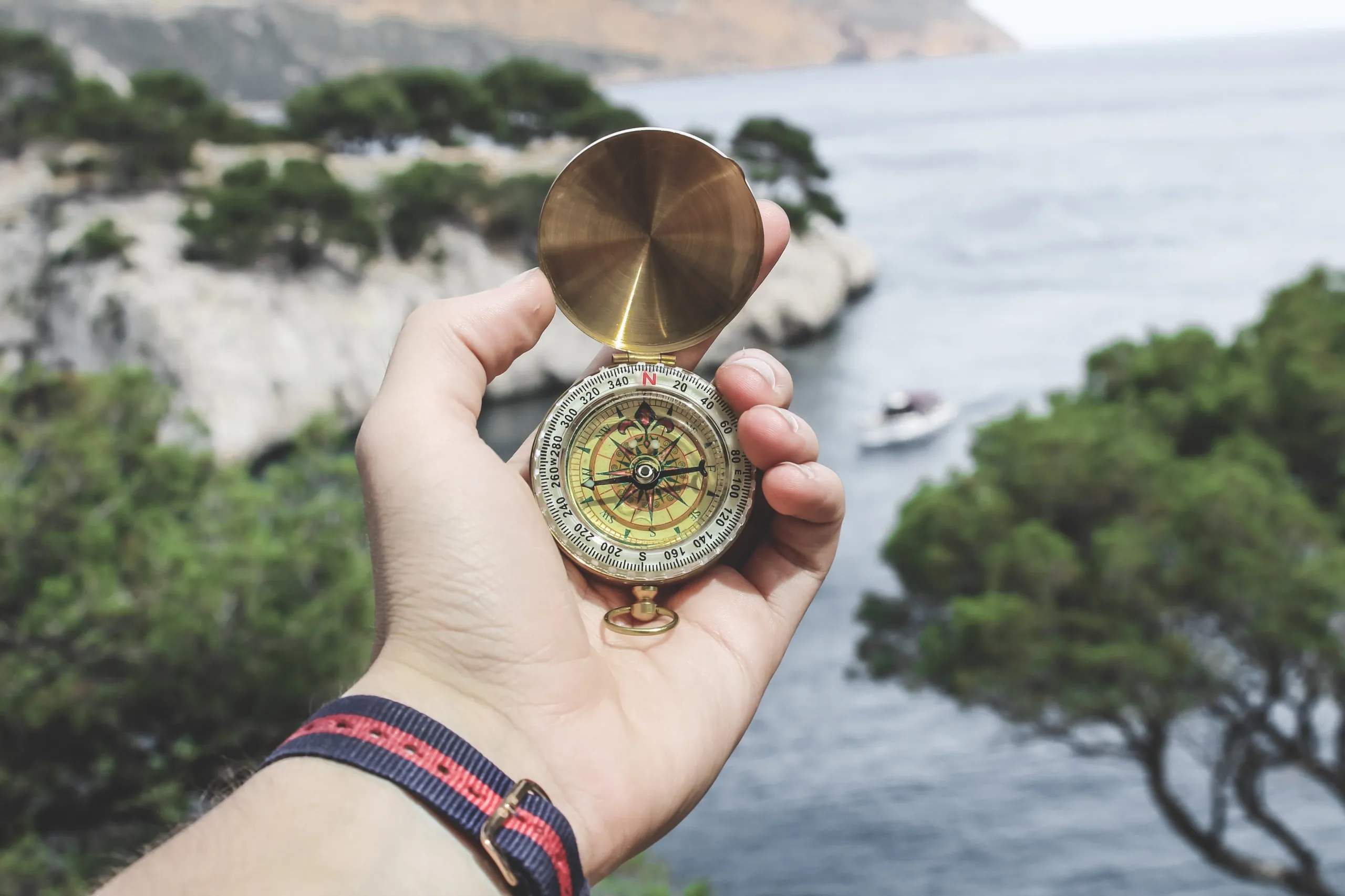 Hand holding an open brass compass with a lake, trees, and a boat in the blurred background.