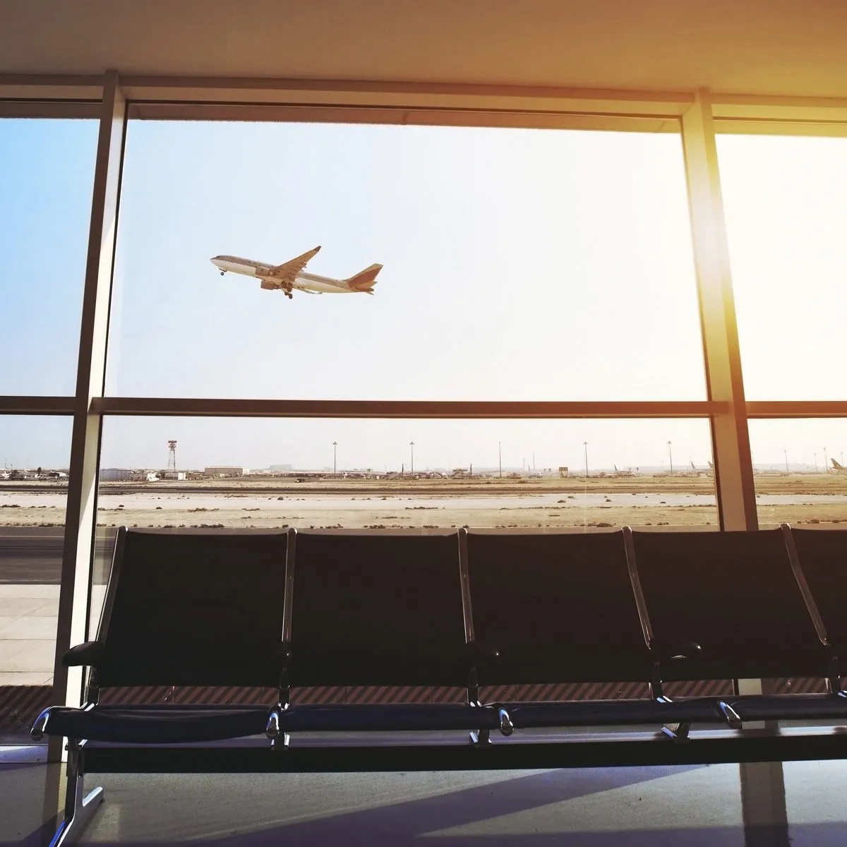 Airplane taking off viewed through large airport terminal windows above empty waiting chairs.
