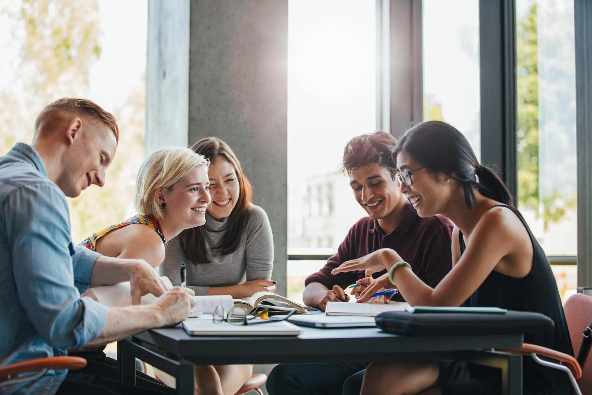Group of five young adults studying together at a table with books and notebooks, smiling and engaged in discussion.