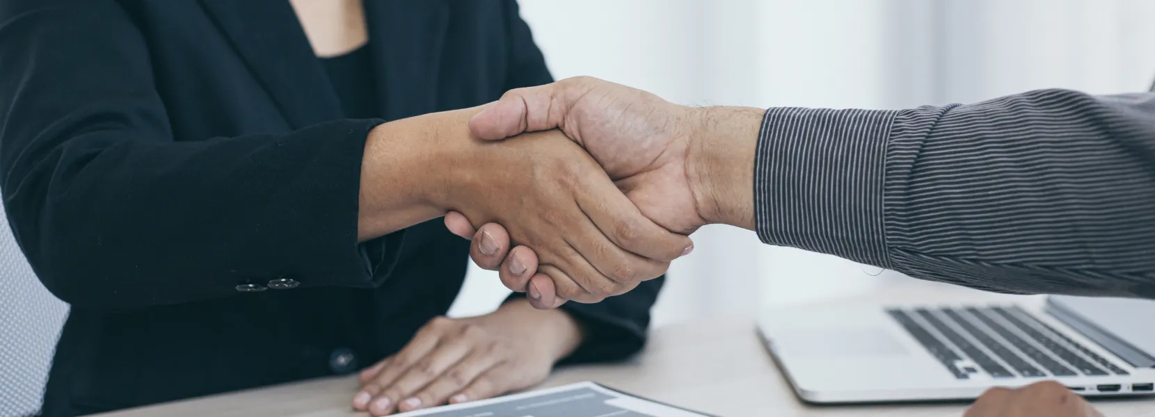 Two people shaking hands over a desk with a laptop and documents, suggesting a business agreement.