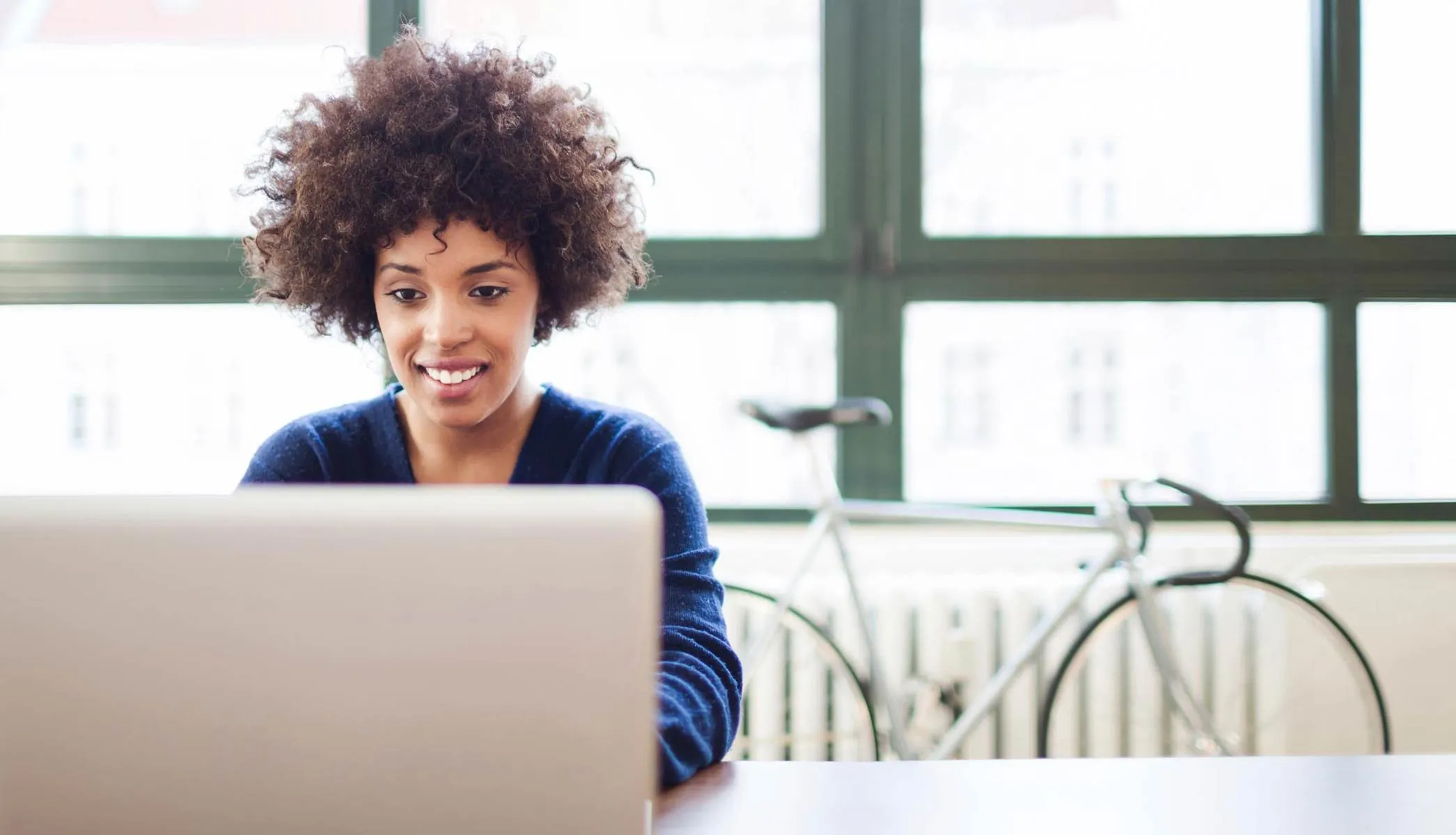 Smiling woman with curly hair using a laptop at a desk with a bicycle and large window in the background.