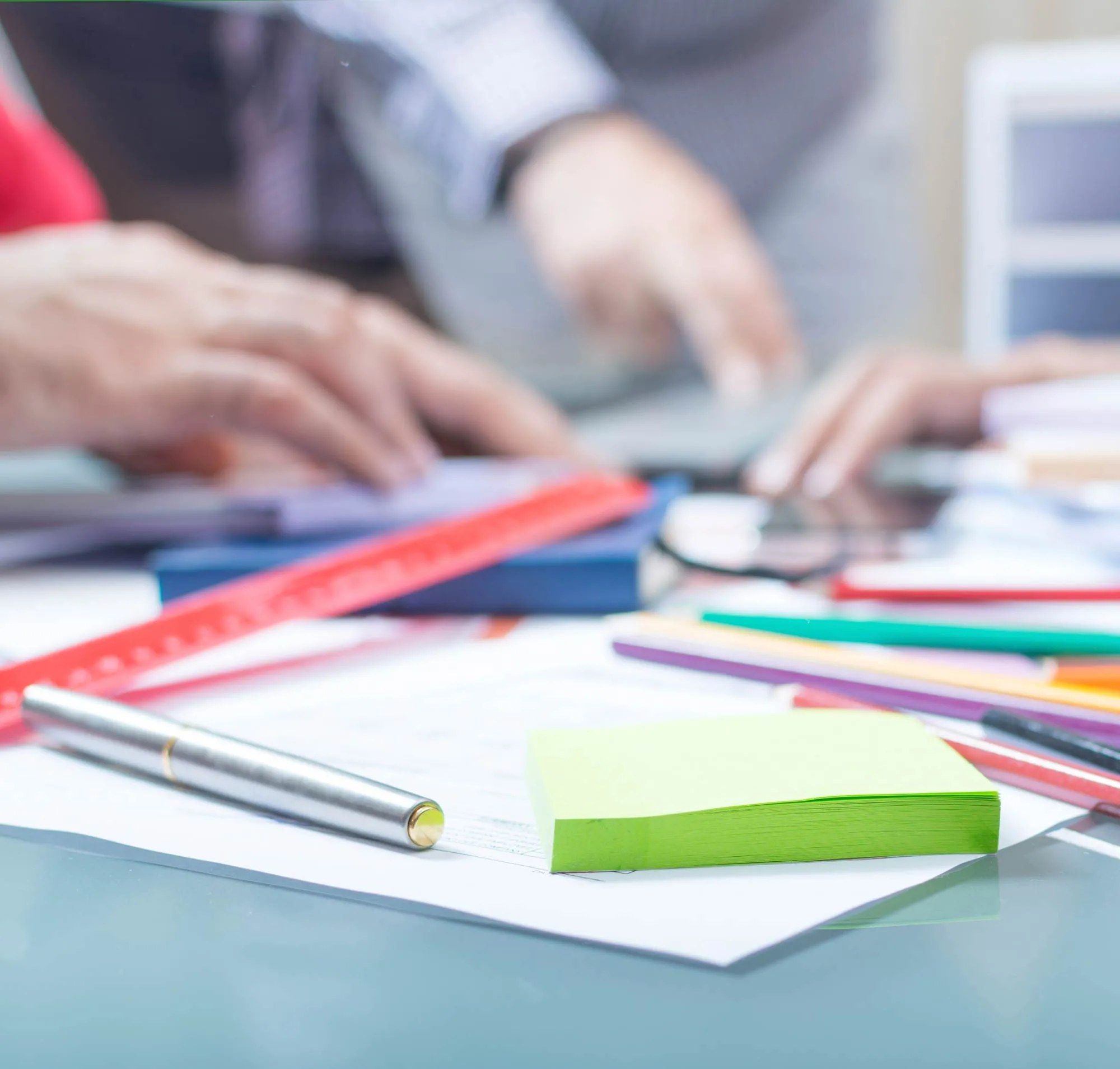 Close-up of a desk with a stack of green sticky notes, a silver pen, colored pencils, and blurred hands working on a laptop in the background.