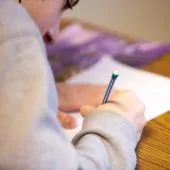 Child wearing glasses writing with a pencil on paper at a wooden table.