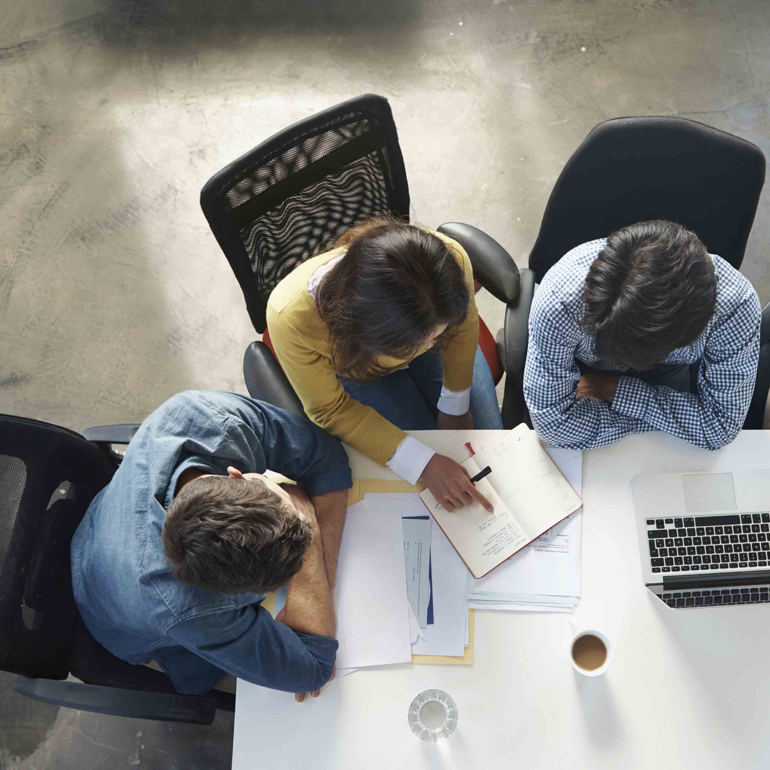 Overhead view of three people sitting at a table with papers, a laptop, a cup of coffee, and a glass of water, discussing notes in a notebook.