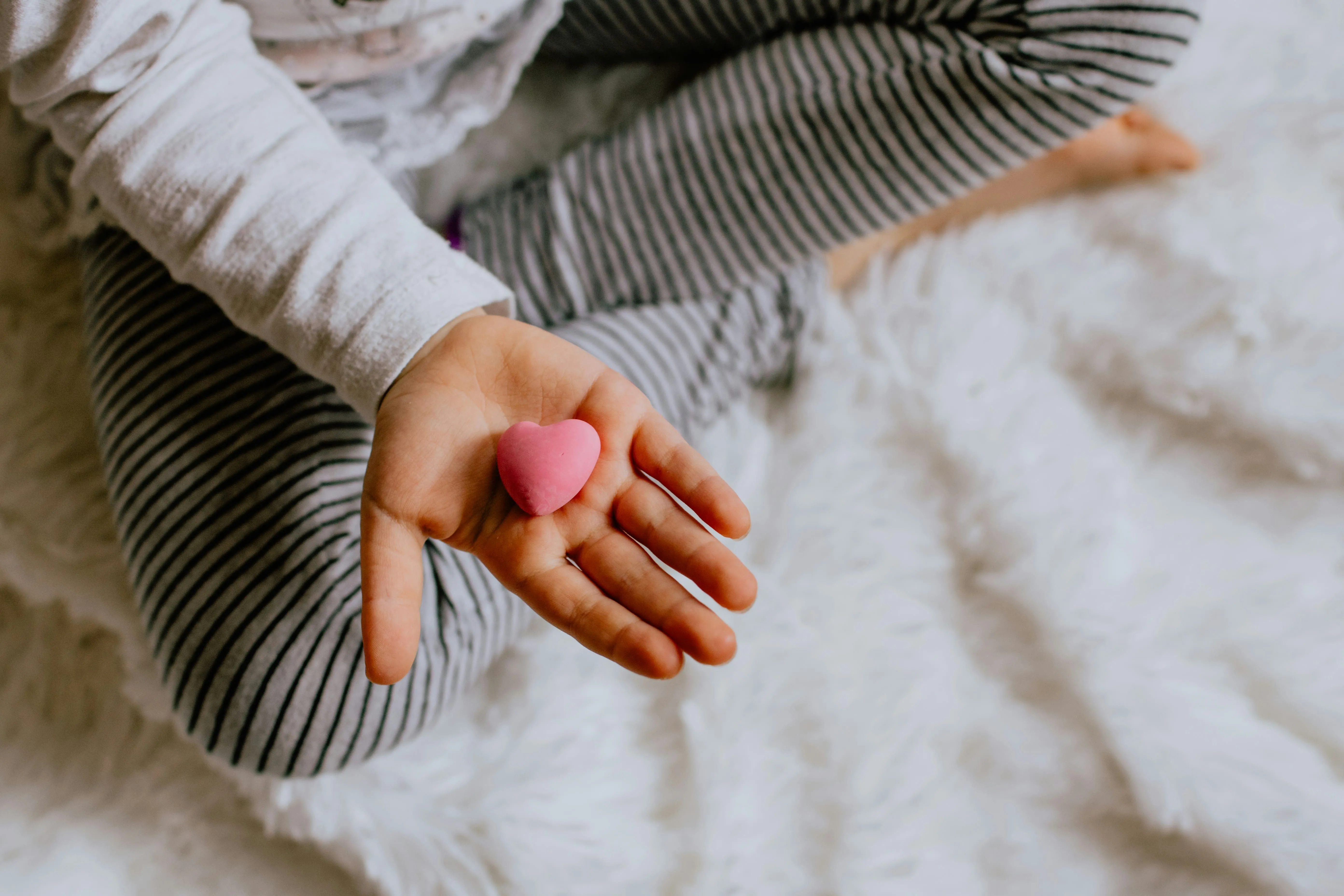 Child sitting cross-legged on a white fuzzy surface, holding a small pink heart-shaped object in an open hand.