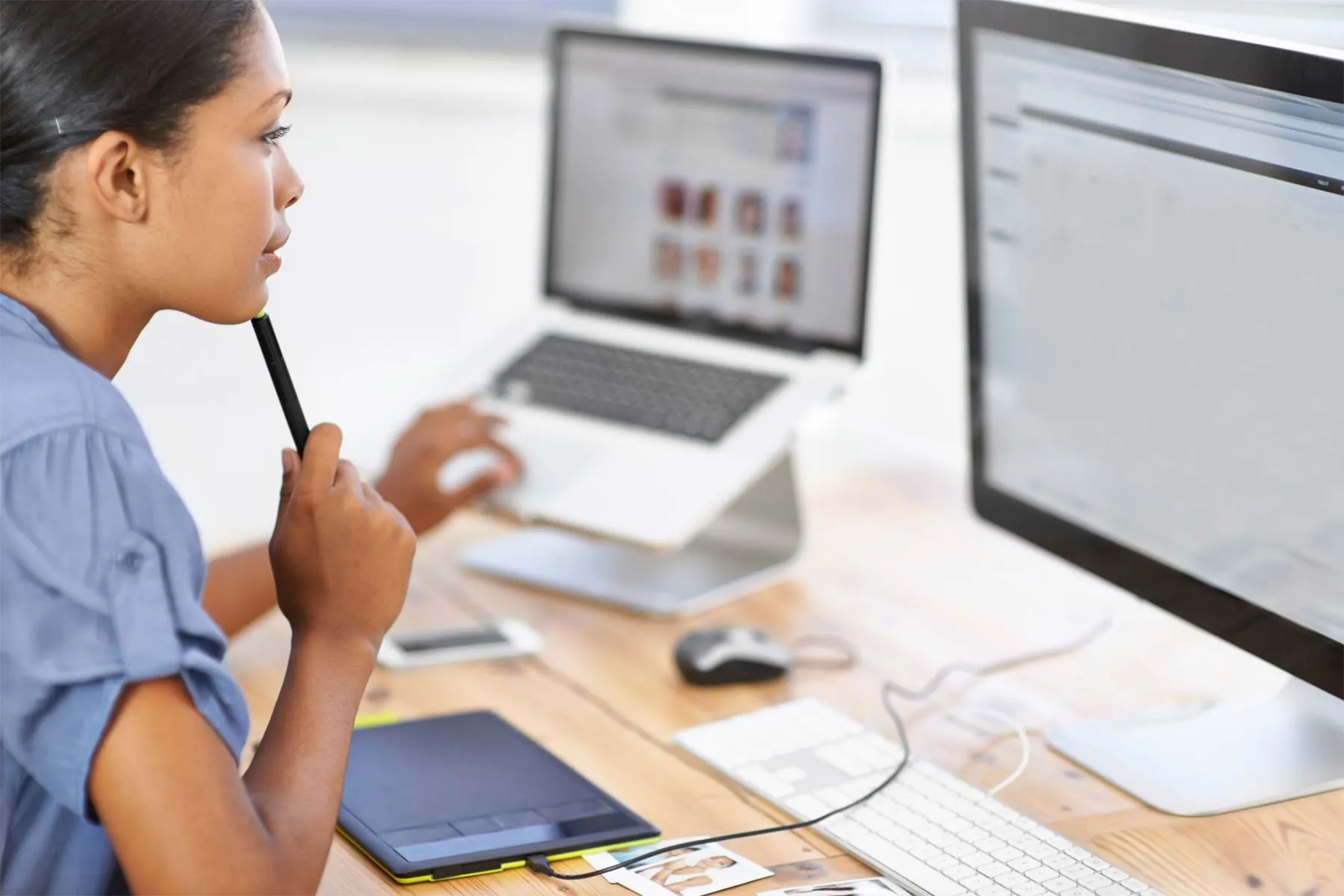 A woman holding a pen to her chin while working on a computer with dual monitors and a laptop on a desk.