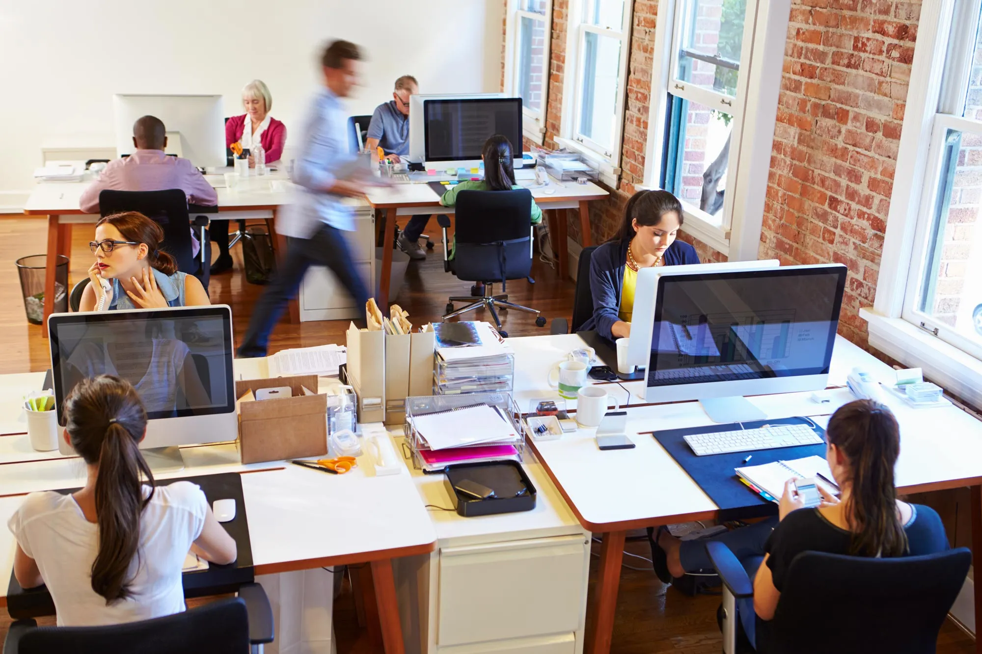 Open office space with diverse coworkers working at desks with computers and office supplies, featuring exposed brick walls and large windows.