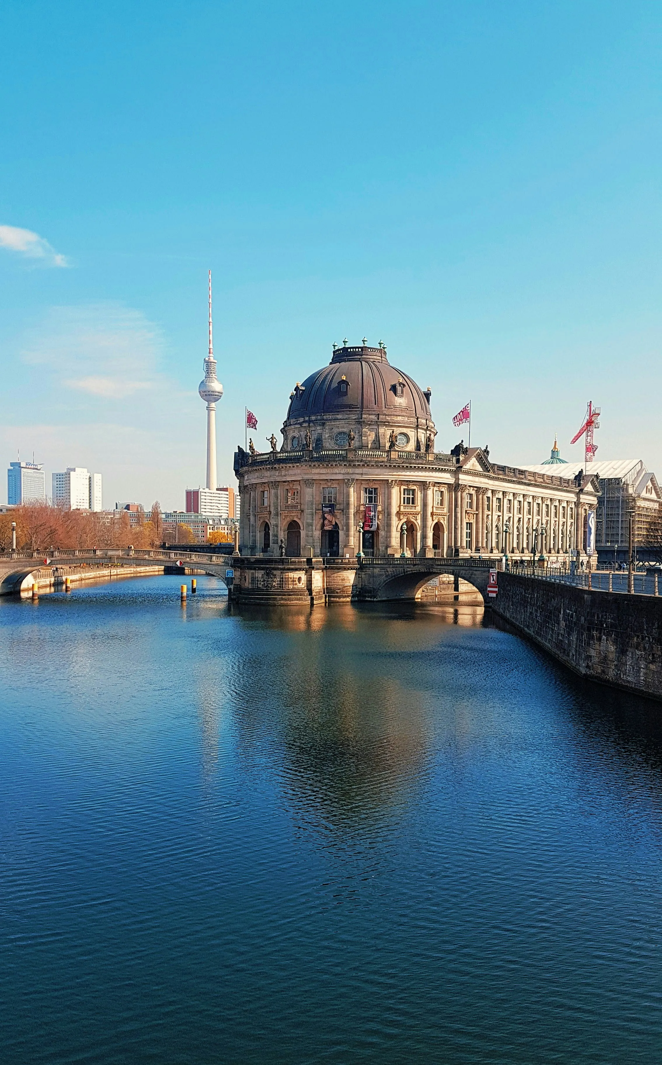 Bode Museum on the Spree River with Berlin TV Tower and city skyline in the background under a clear blue sky.