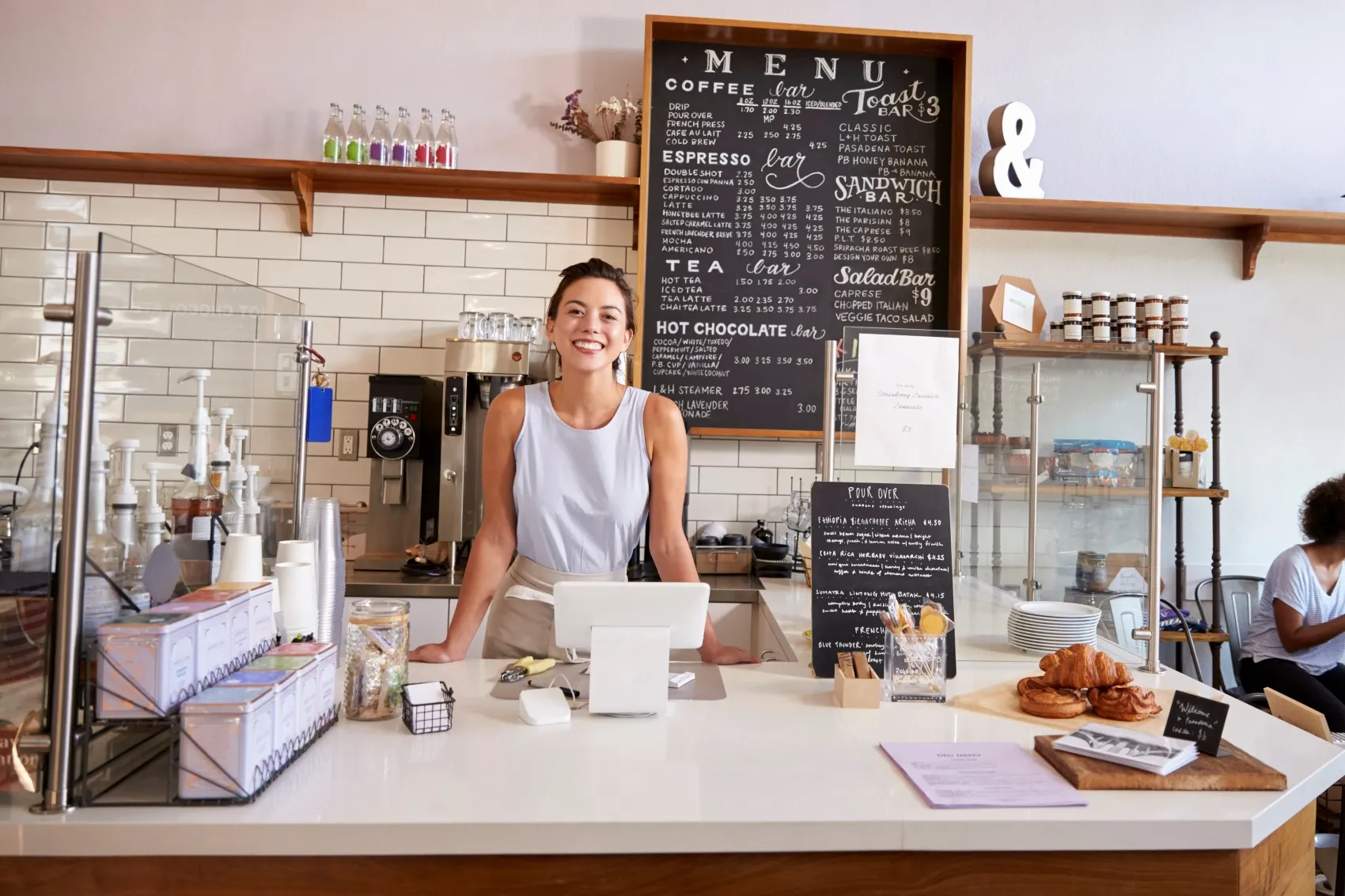 Smiling barista standing behind a café counter with a menu board, pastries, and coffee equipment in the background.