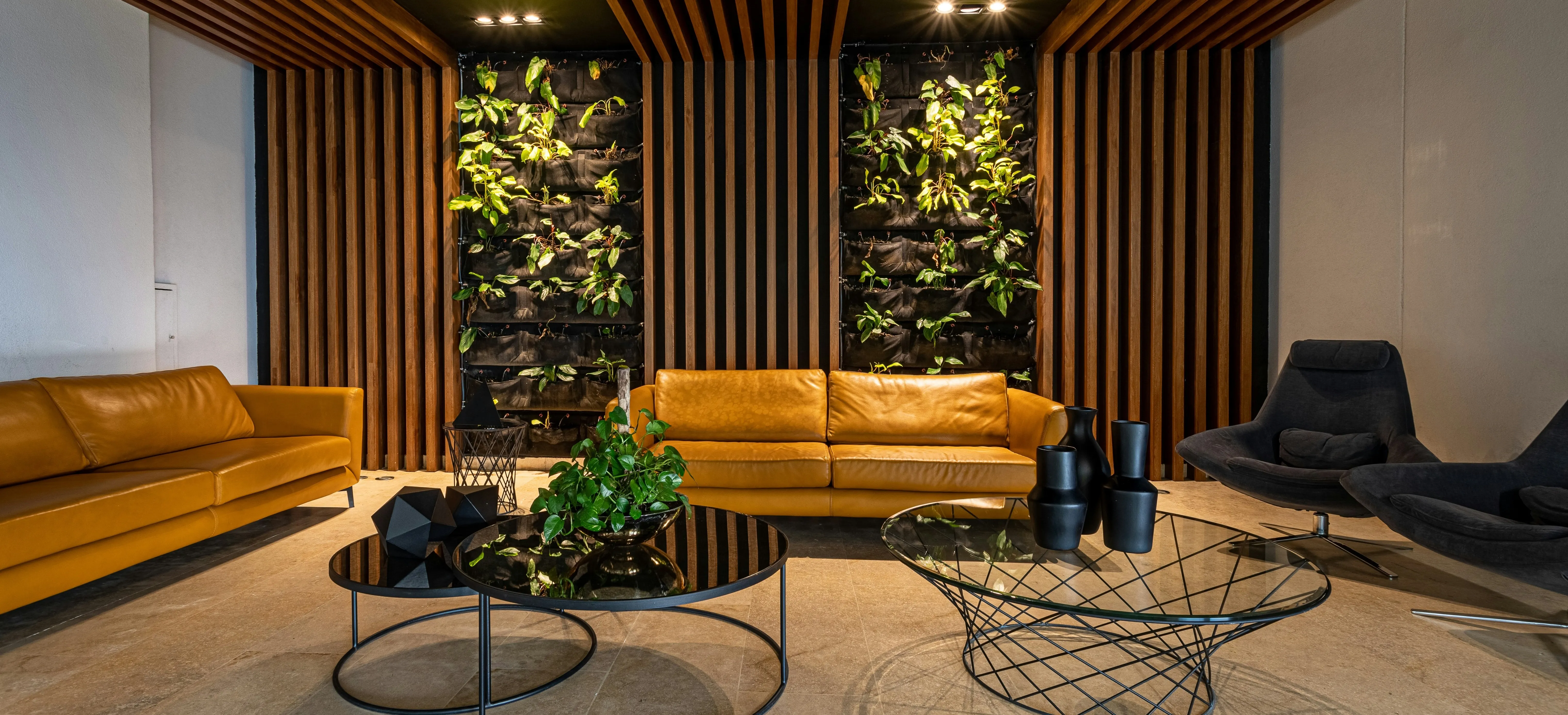Modern lounge area with two mustard yellow sofas, two black mesh coffee tables with decorative plants and black vases, accented by wood slat walls and vertical green plant panels.