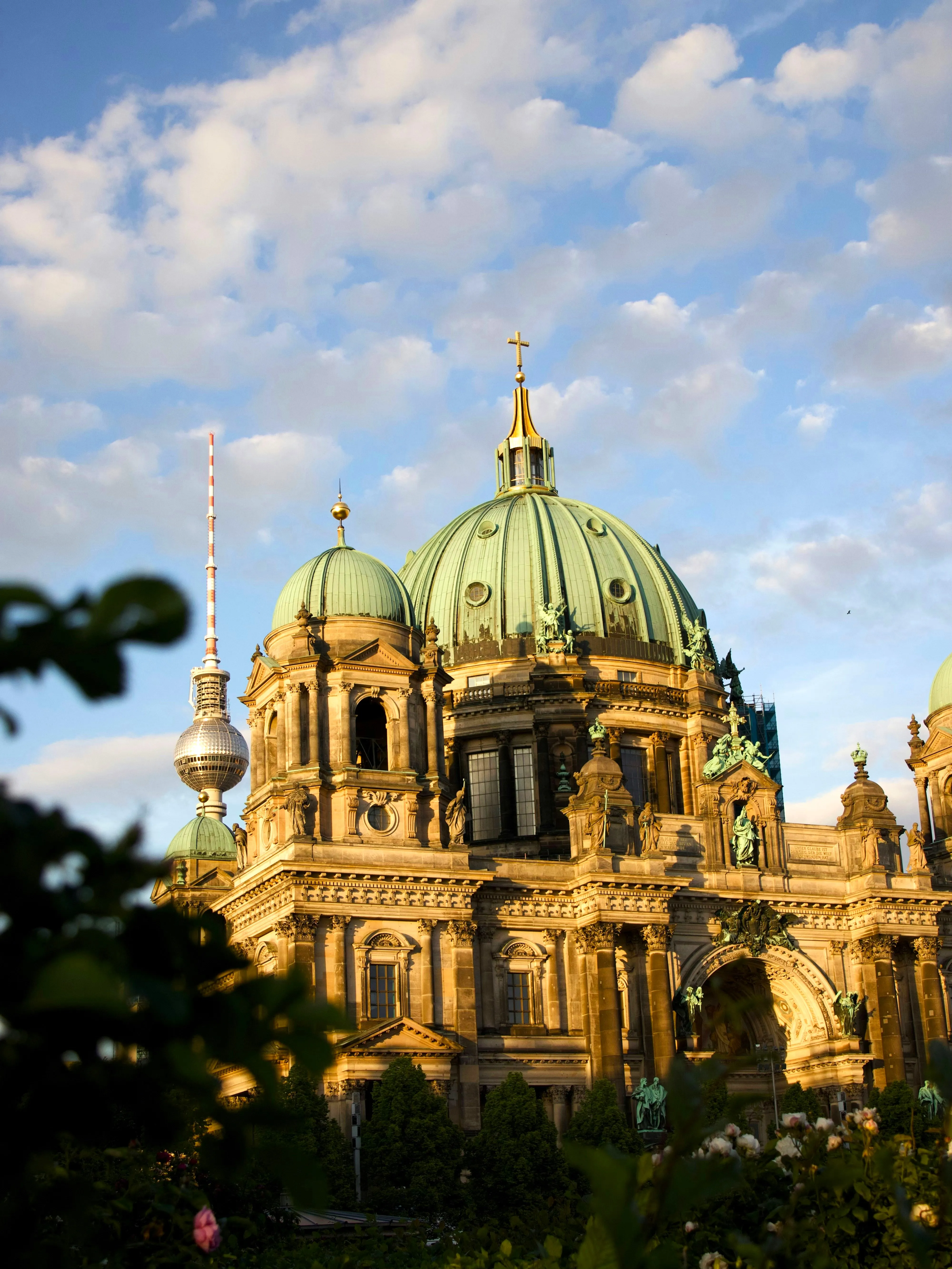 Berlin Cathedral with green domes illuminated by golden sunlight against a partly cloudy blue sky, with TV tower in the background.