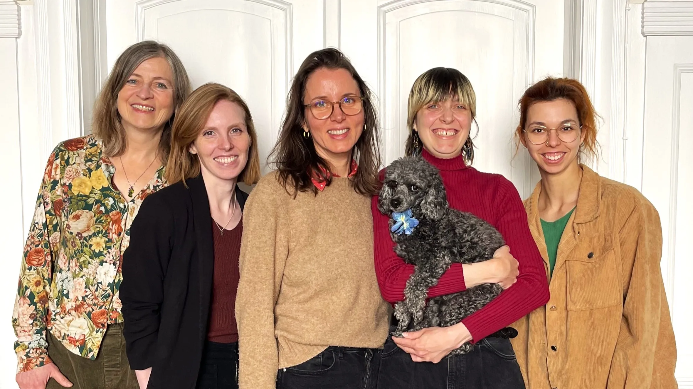 Five women standing and smiling, one holding a small gray poodle.