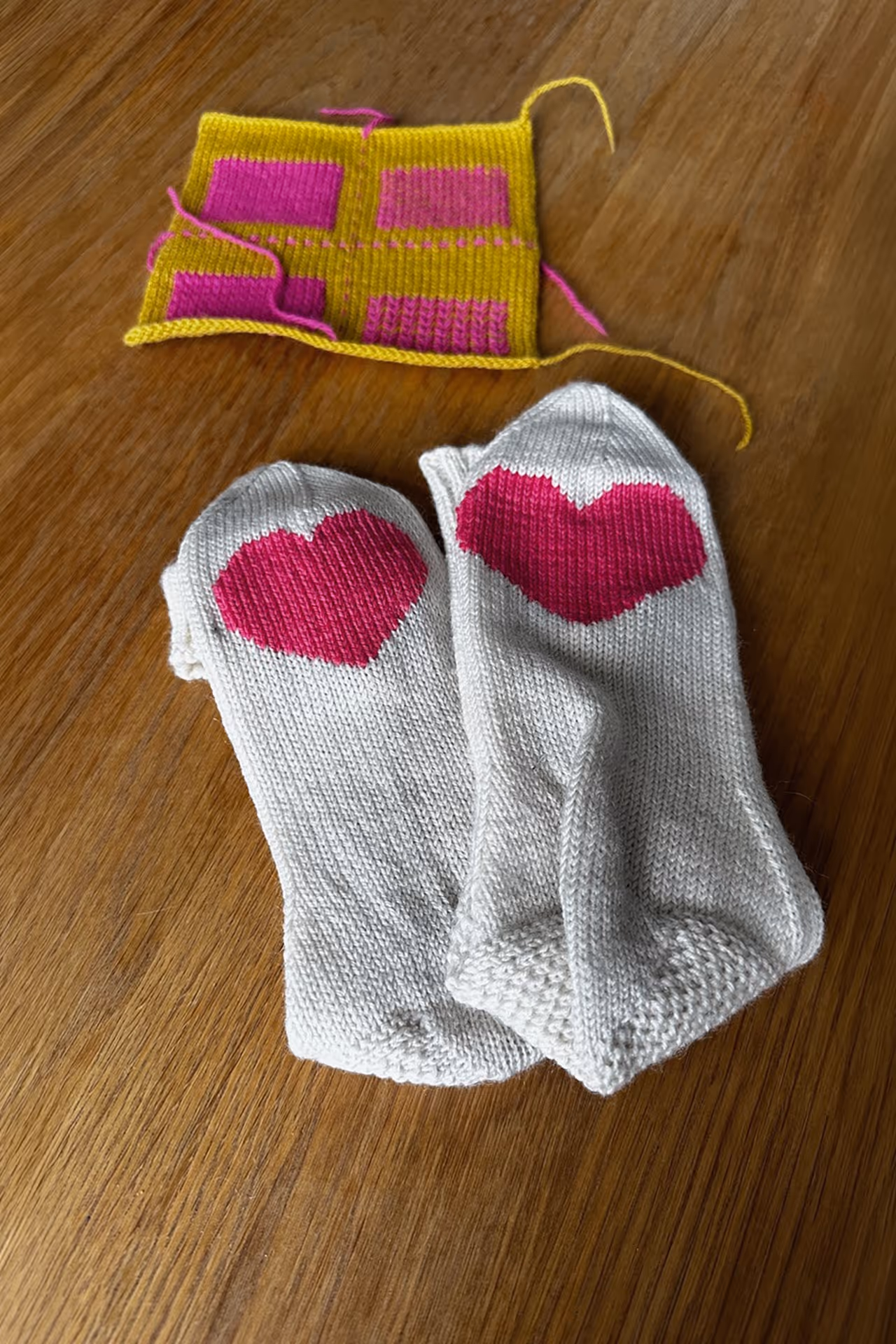 Gestrickte Socken mit pinken Herzmotiven neben einem Beispielstück für Stopftechniken auf Holzuntergrund.