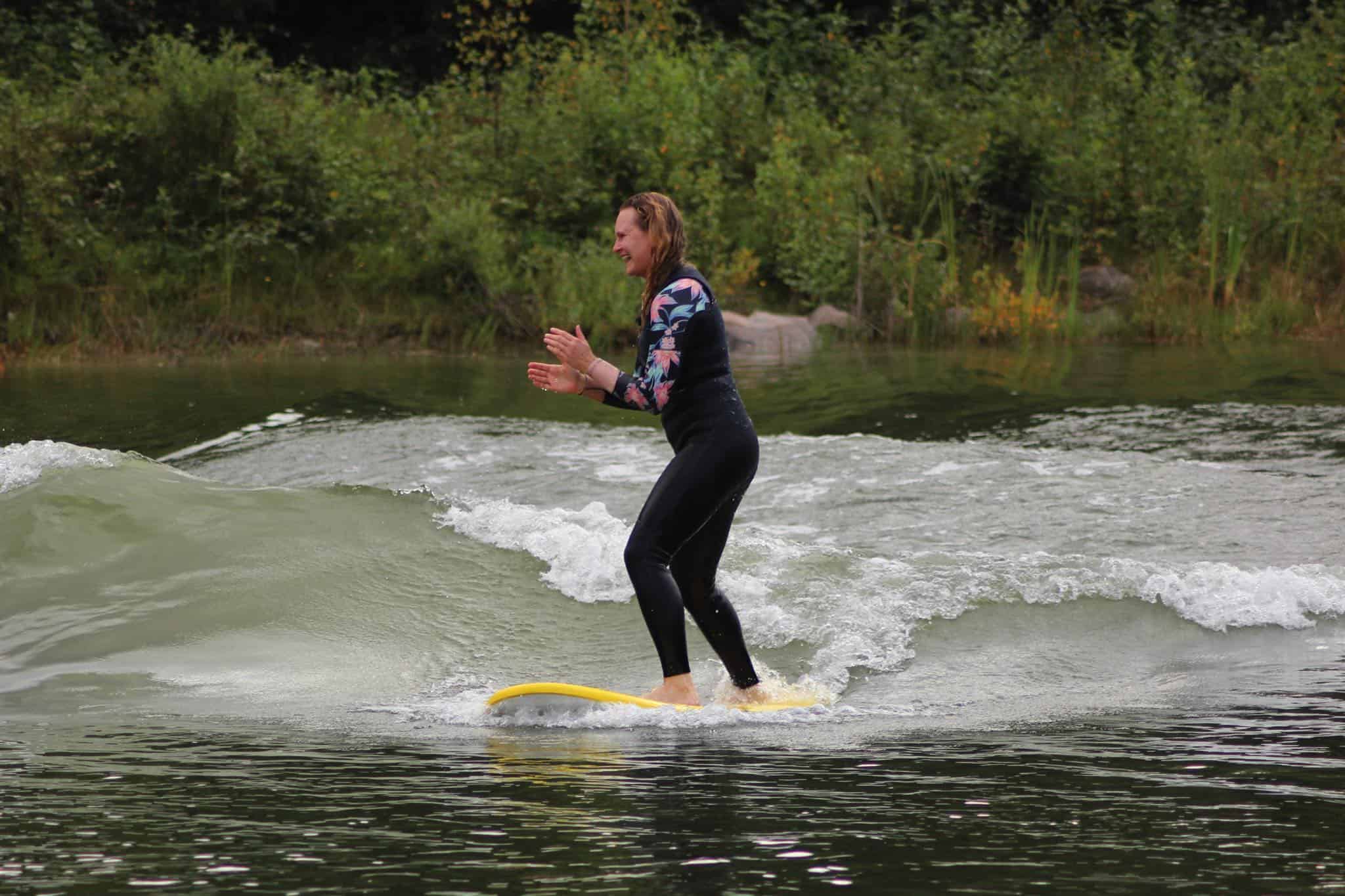 girl on surfboard surfing in lake clapping hands in black and colourful wetsuit. Image has green bushes in background