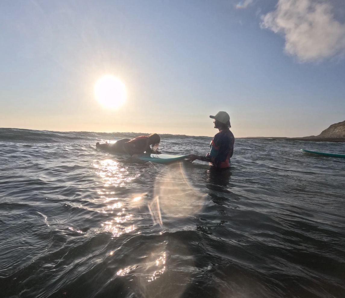 surf coach standing in ocean, beginner surfing laying on surfboard waiting for wave, sun setting in the background