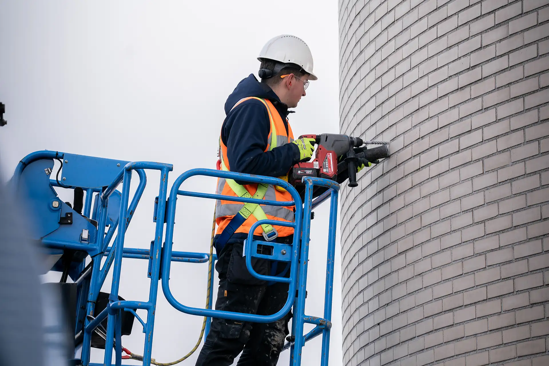 Bouwvakker in veiligheidshelm en oranje veiligheidsvest gebruikt een boor op een lichtgrijze bakstenen muur vanaf een blauwe hoogwerker.