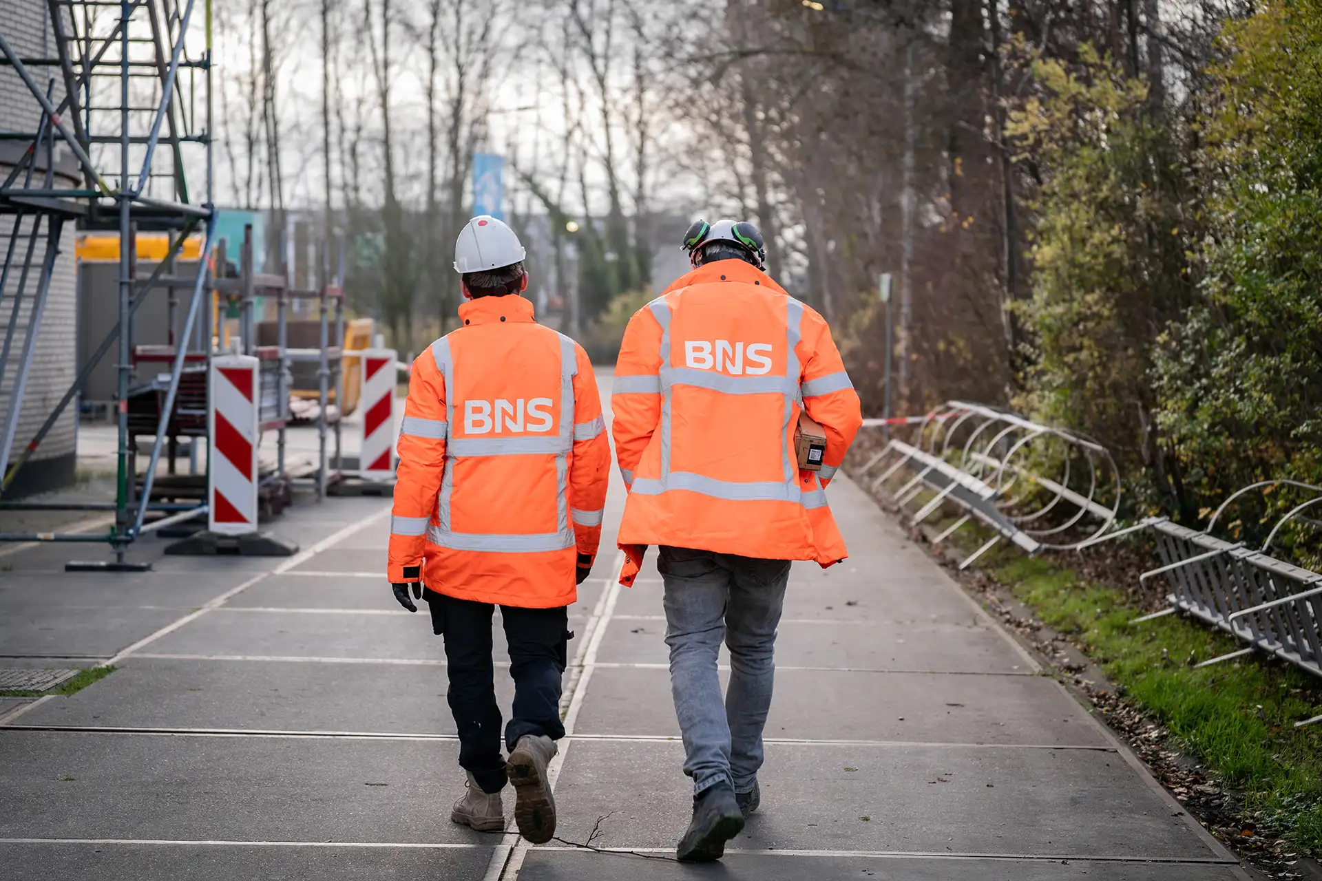 Twee bouwvakkers in oranje veiligheidsjassen met BNS-logo lopen op betonnen vloer nabij bouwmateriaal en struiken.