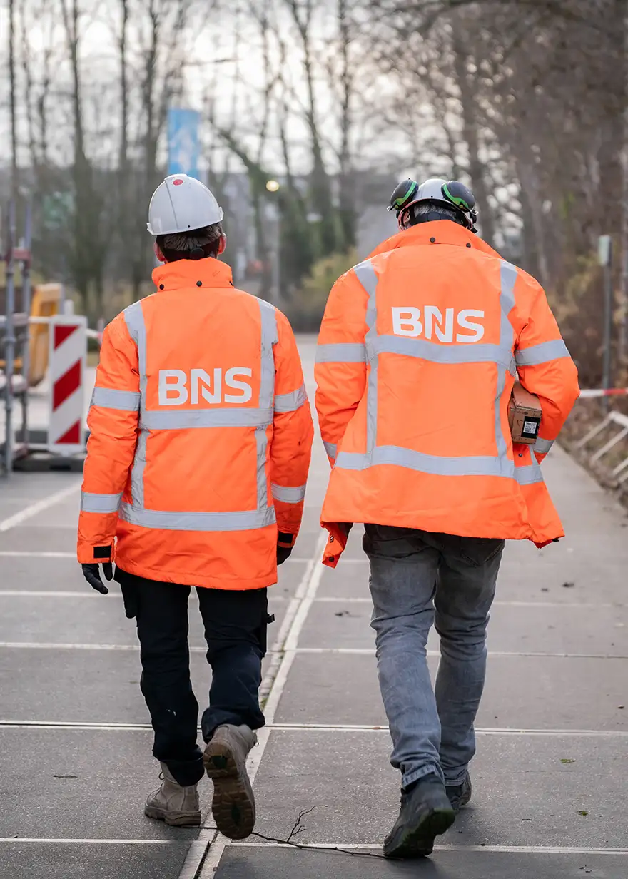 Twee bouwvakkers in oranje veiligheidsjassen met 'BNS' op de rug lopen op een bouwplaats.