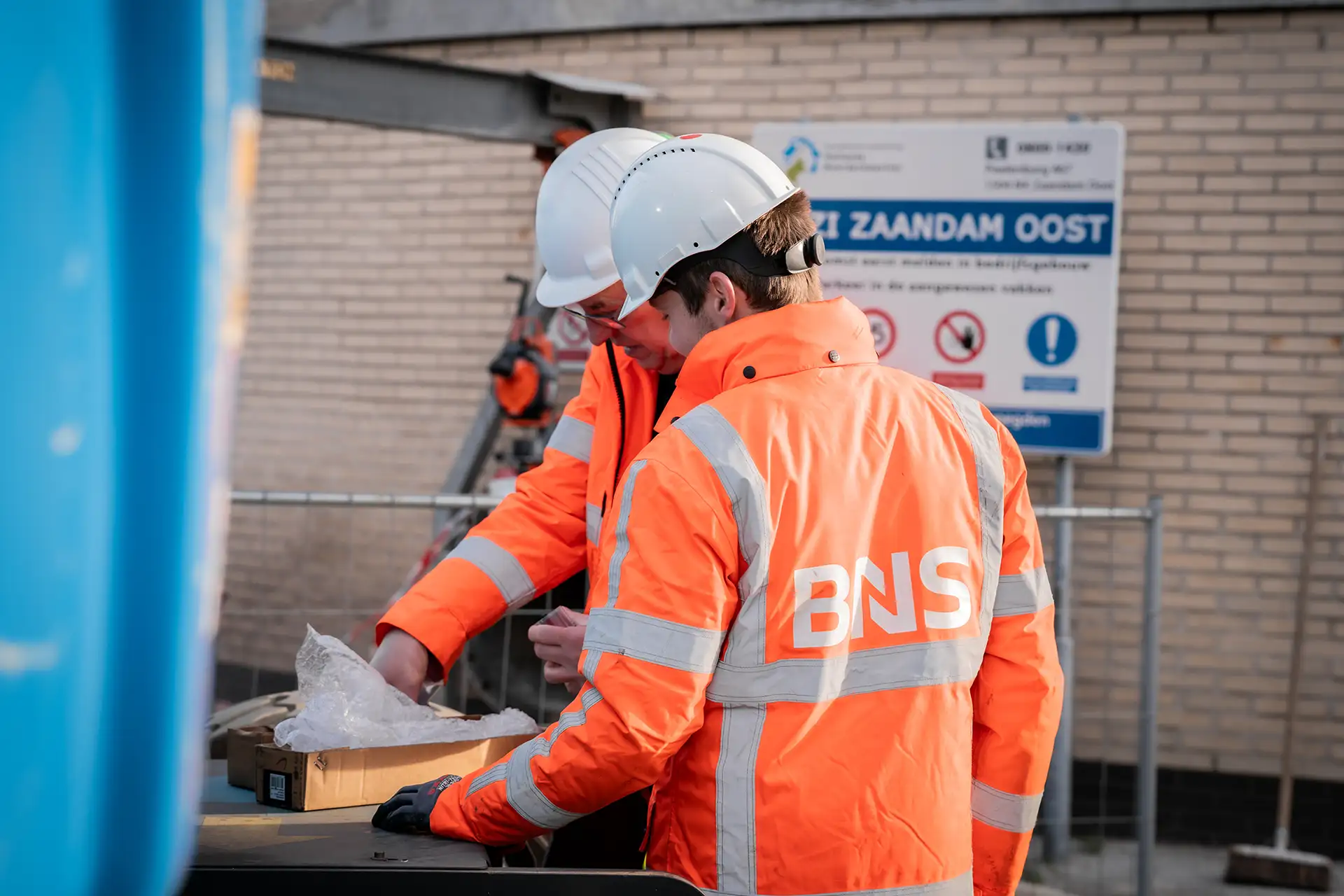 Twee bouwvakkers in oranje jassen en witte helmen bekijken een doos op een bouwplaats met een bord op de achtergrond.