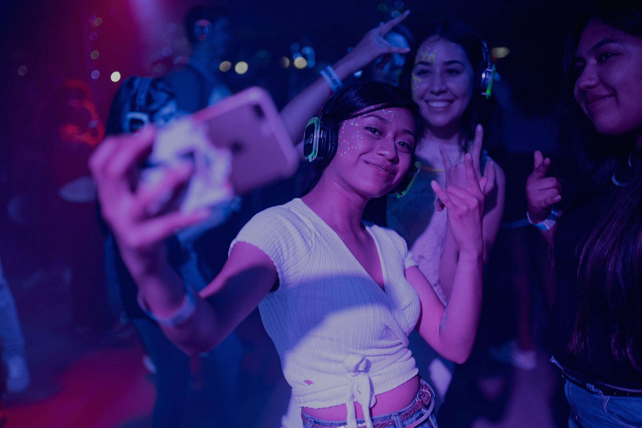 Young women wearing headphones and face paint taking a selfie at a party with purple and blue lighting.
