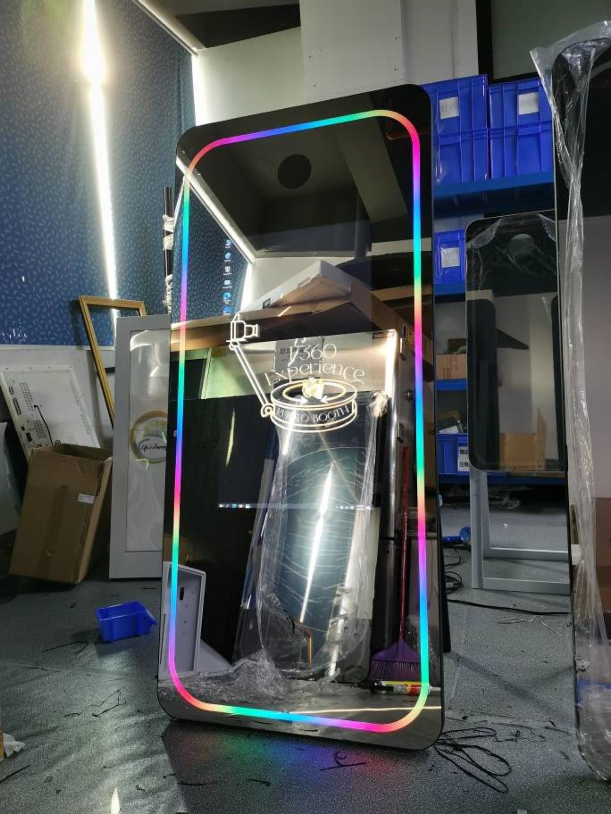 Tall rectangular mirror with a colorful LED border displaying 'F360 Experience Photo Booth' logo, placed in a cluttered room.