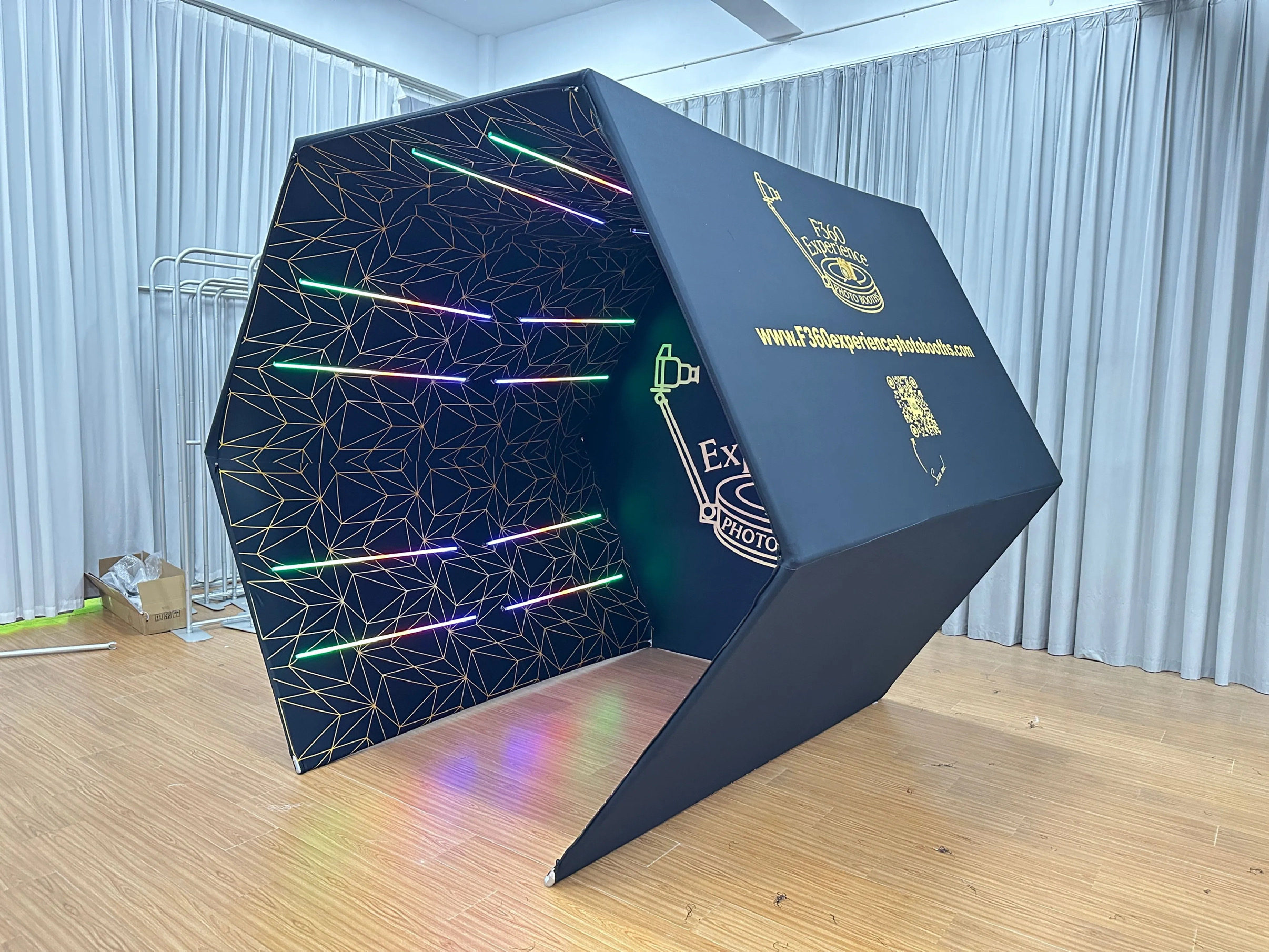 Modern hexagonal photo booth with multicolored LED lights inside, placed on wooden floor with white curtains in background.
