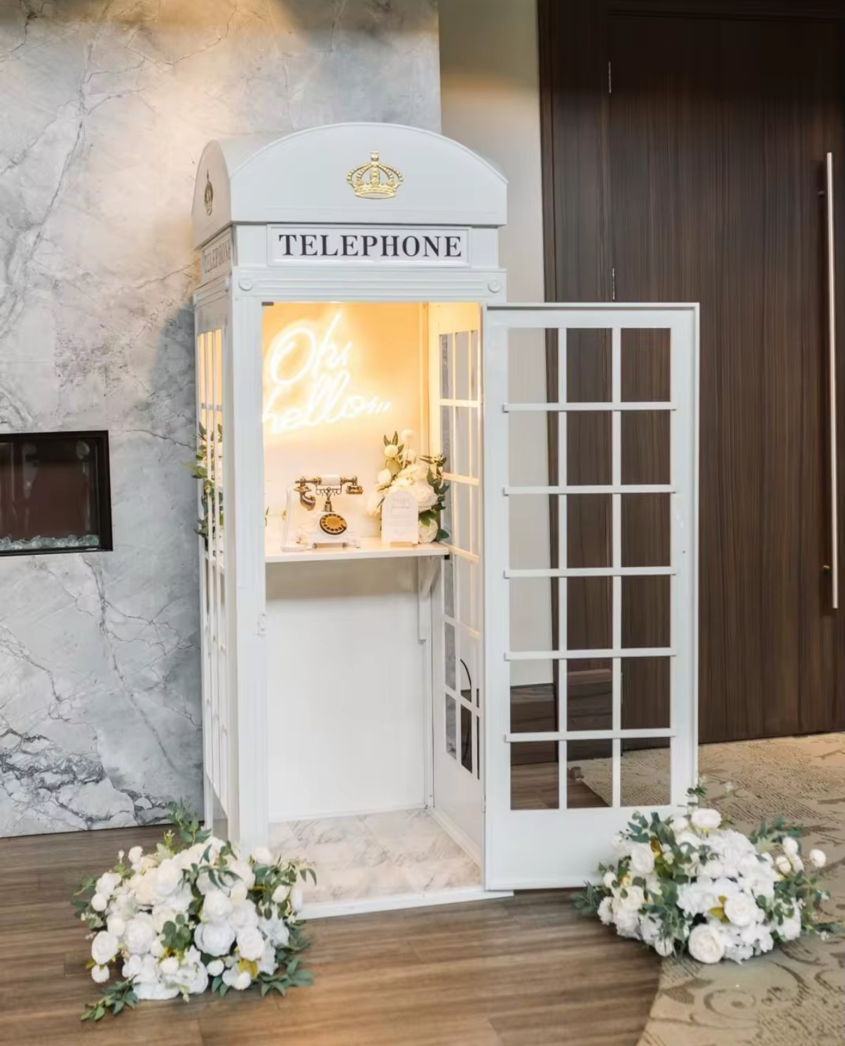 White decorative telephone booth with open door, inside a vintage rotary phone and illuminated neon sign reading 'Oh hello', surrounded by white floral arrangements.