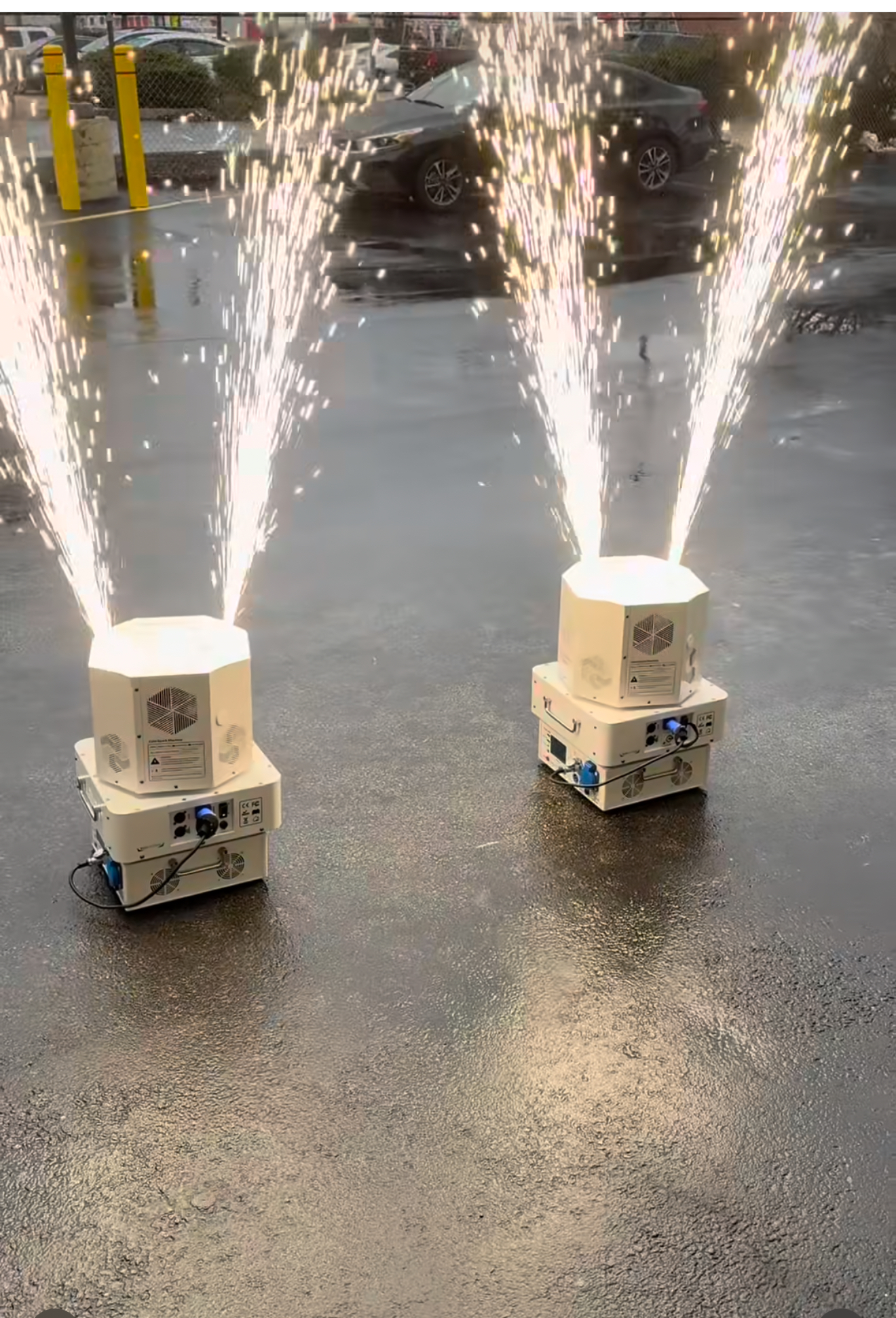 Two white machines on wet pavement shooting bright sparkler-style fireworks upward.
