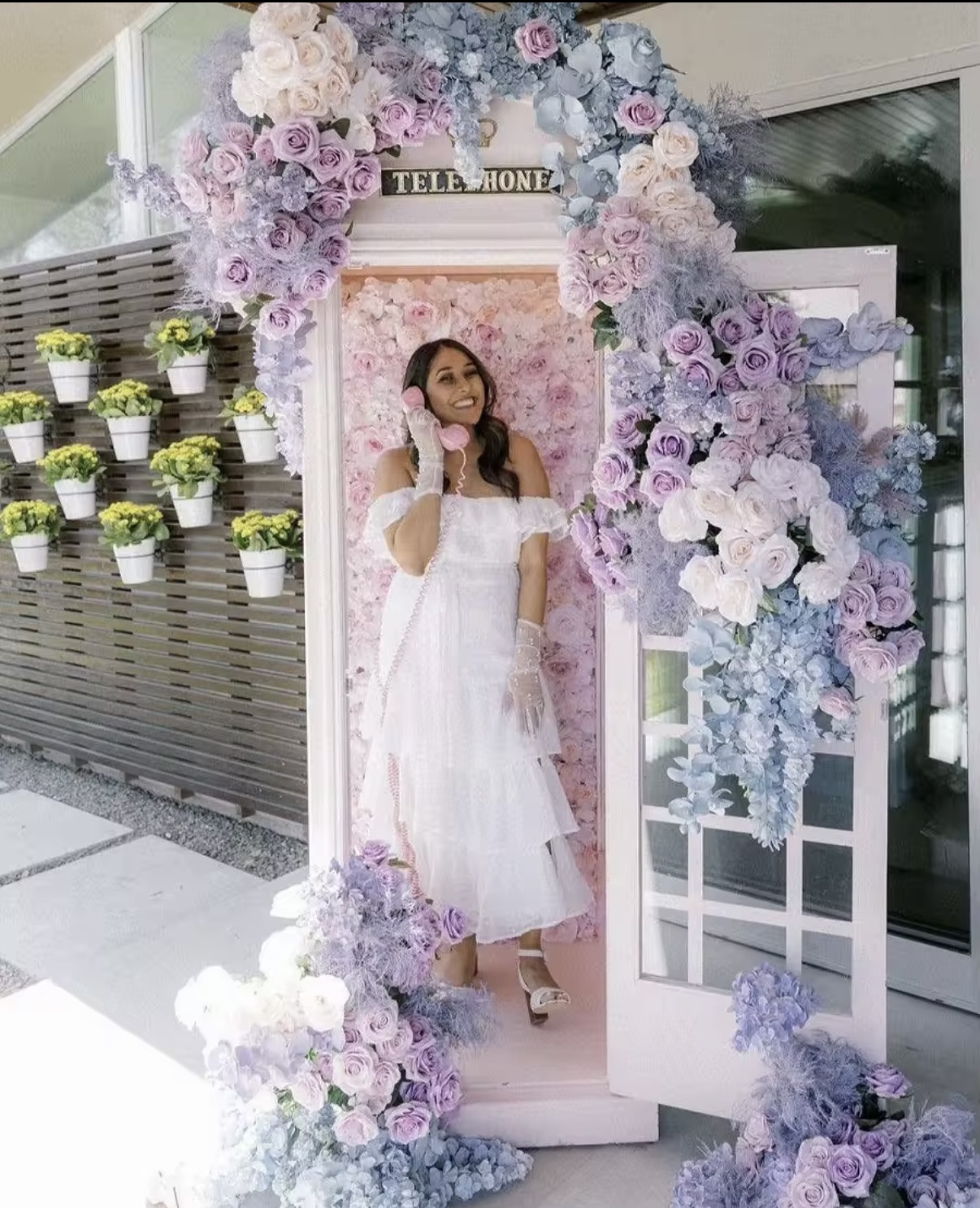 Woman in a white dress smiling inside a floral-decorated vintage telephone booth holding a pink phone receiver.