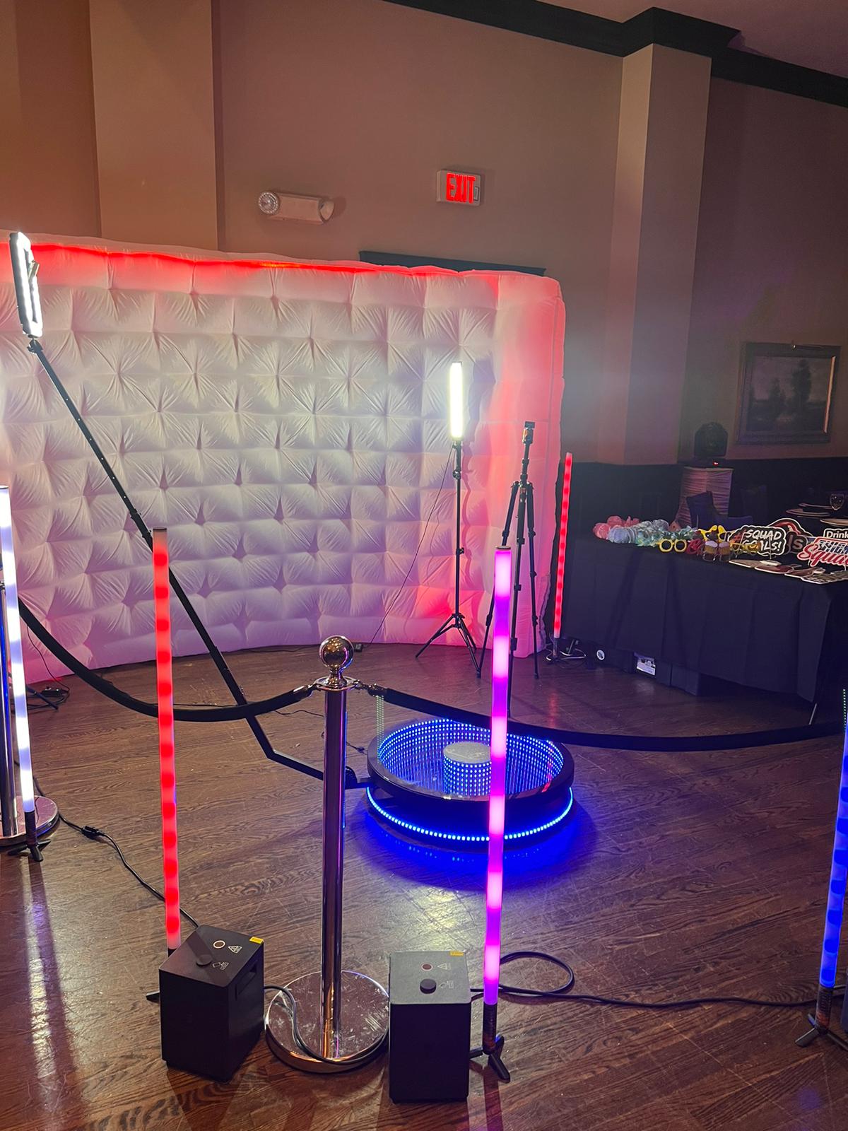 Photo booth setup with LED light tubes in pink, blue, and white, a circular platform with infinity LED lights, and a white cushioned backdrop with red lighting edges, inside a room with wooden flooring.